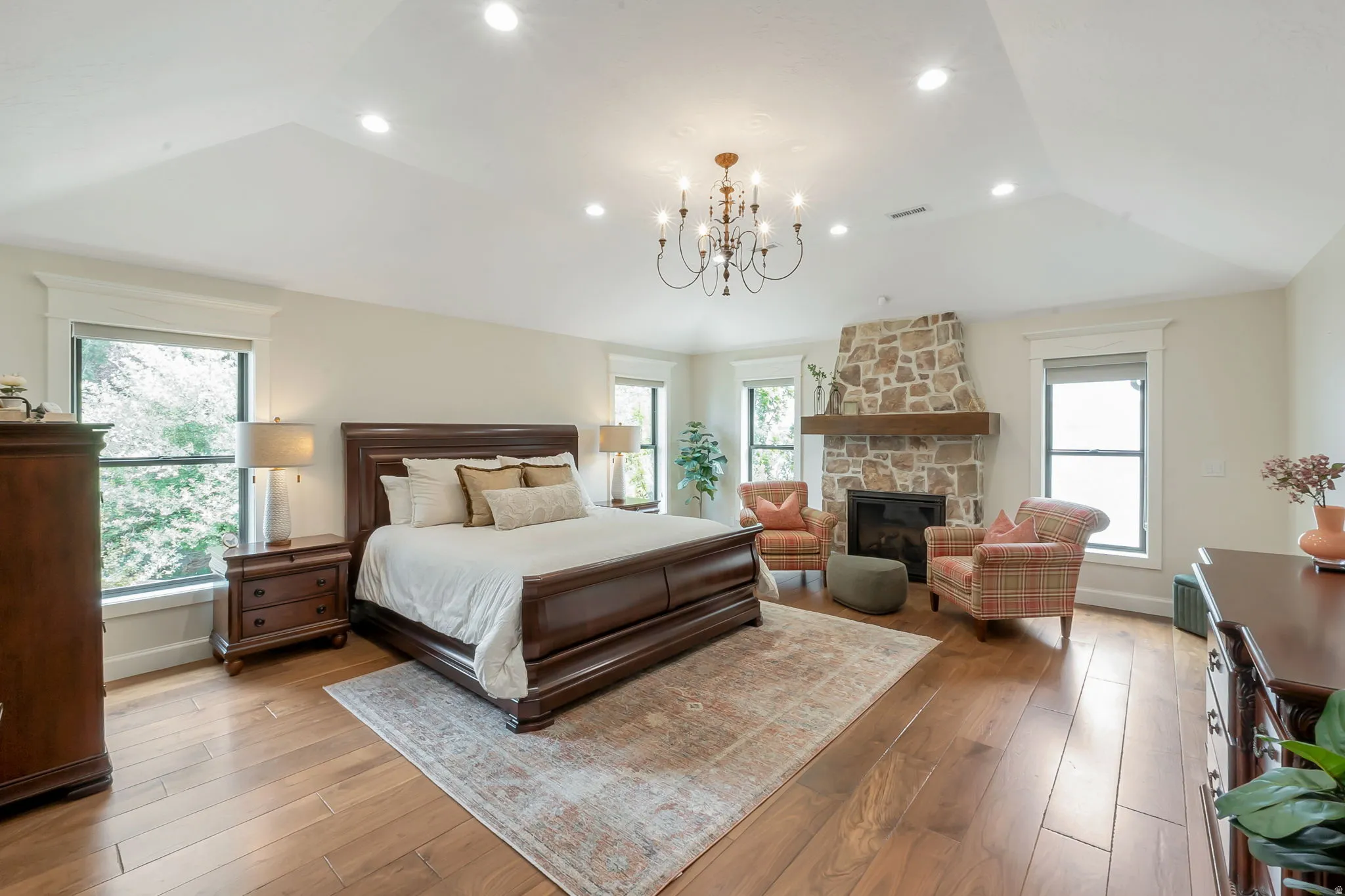 Spacious room featuring wood-finish flooring, a vaulted ceiling with recessed lighting and a chandelier, large windows with white trim, and a stone-faced fireplace with a wood mantel