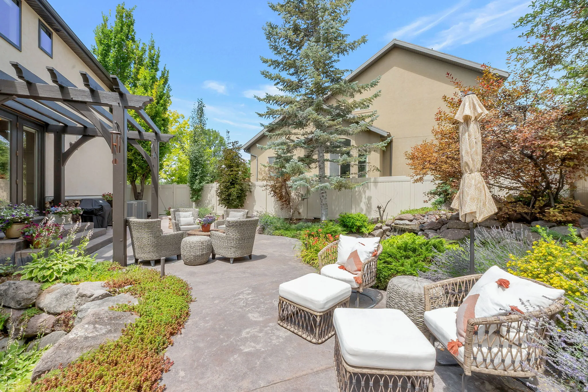 Expansive hardscaped patio area featuring a wooden pergola, lush rock garden landscaping, and mature trees
