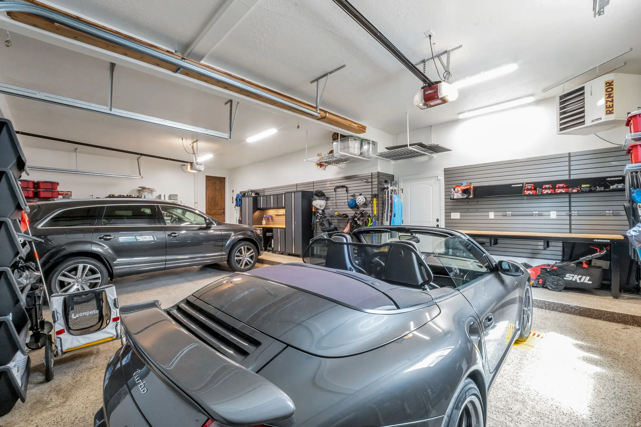 Spacious garage featuring ample overhead lighting, organized slatwall storage systems, built-in cabinetry, and a dedicated workspace with a countertop
