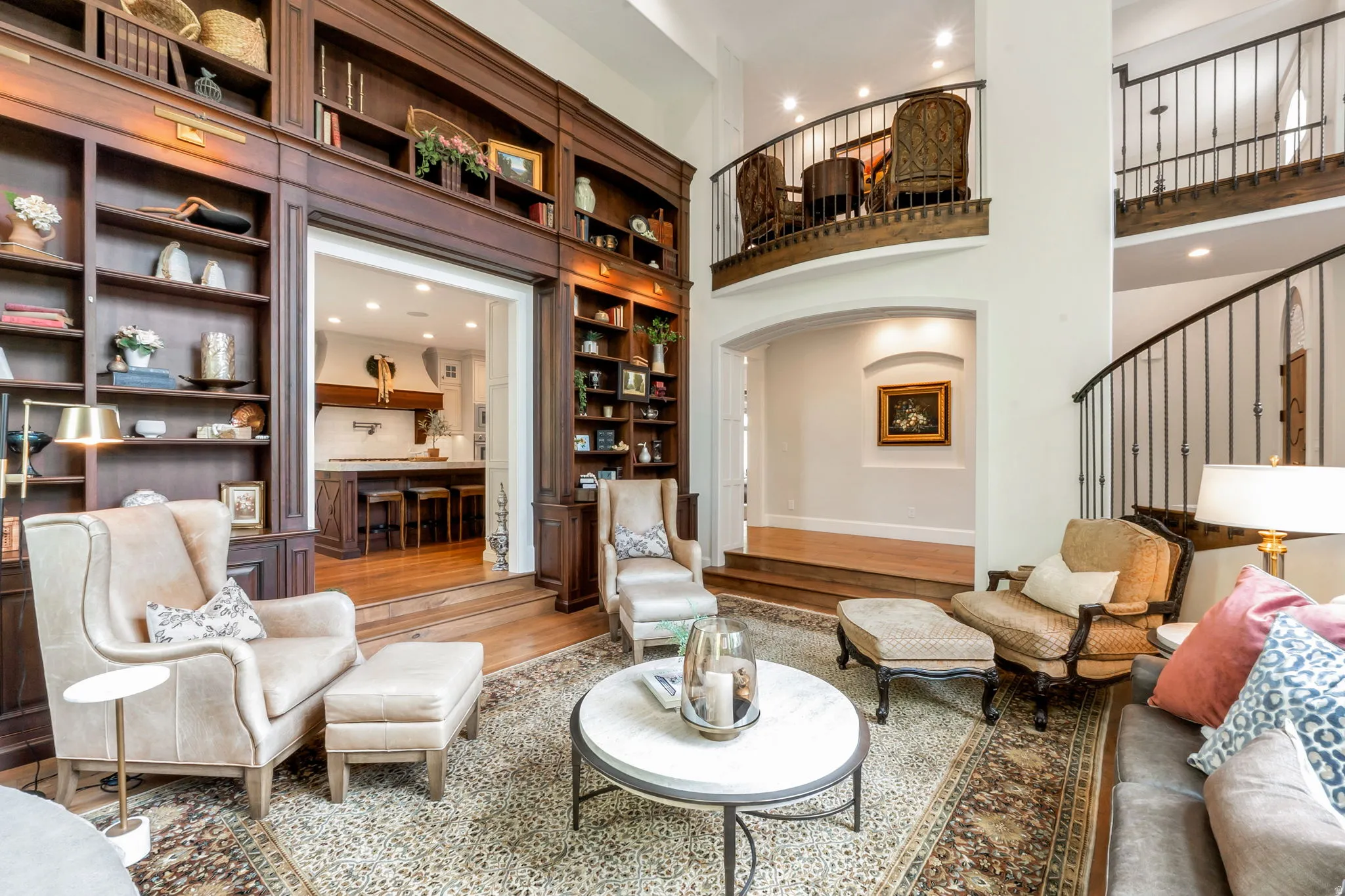 Two-story living area featuring extensive built-in wooden shelving, an open archway to the kitchen, a curved staircase with wrought iron balusters, and a second-story balcony
