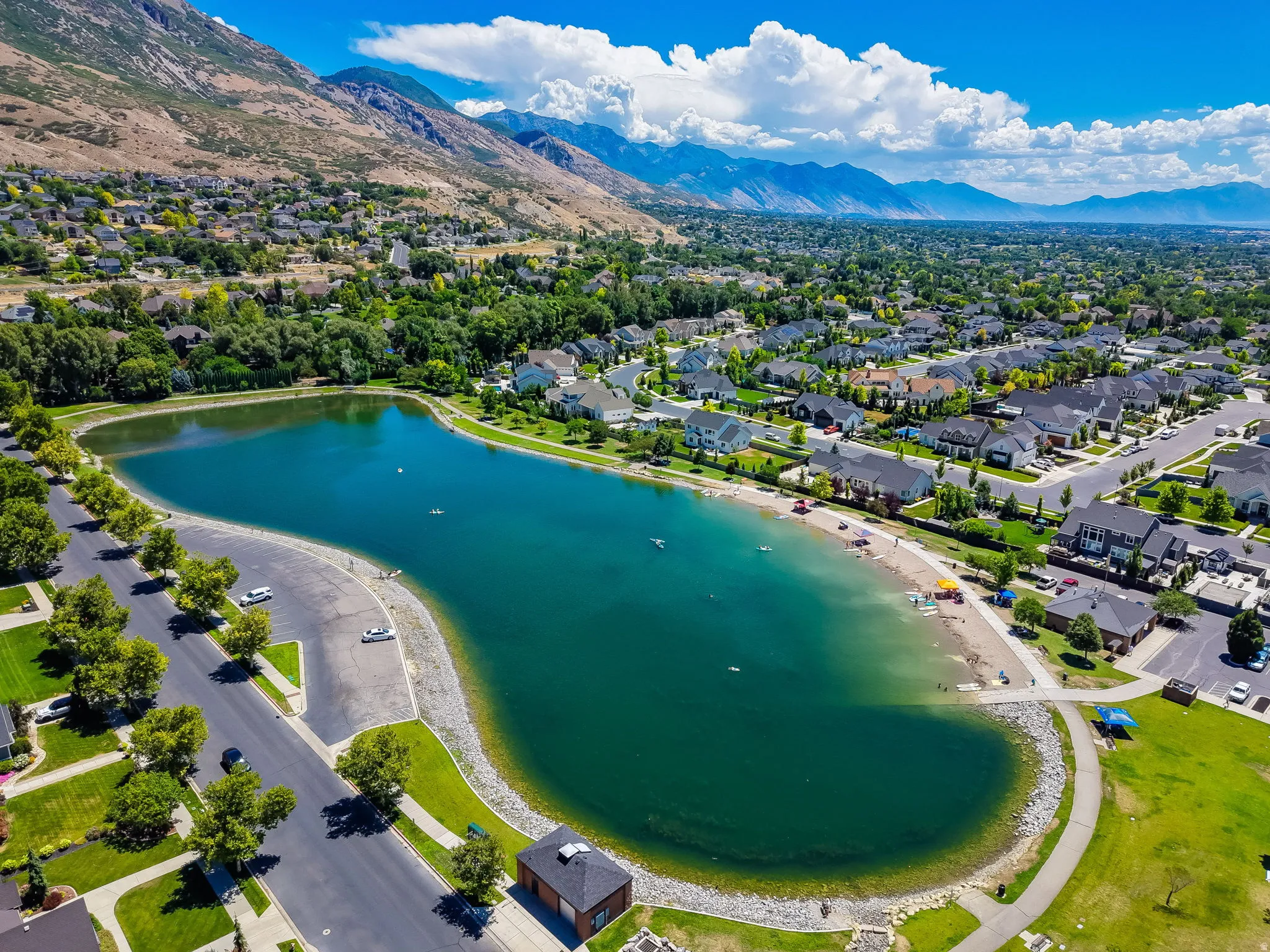 Expansive community lake featuring a sandy beach area, surrounding green spaces, and paved walking paths