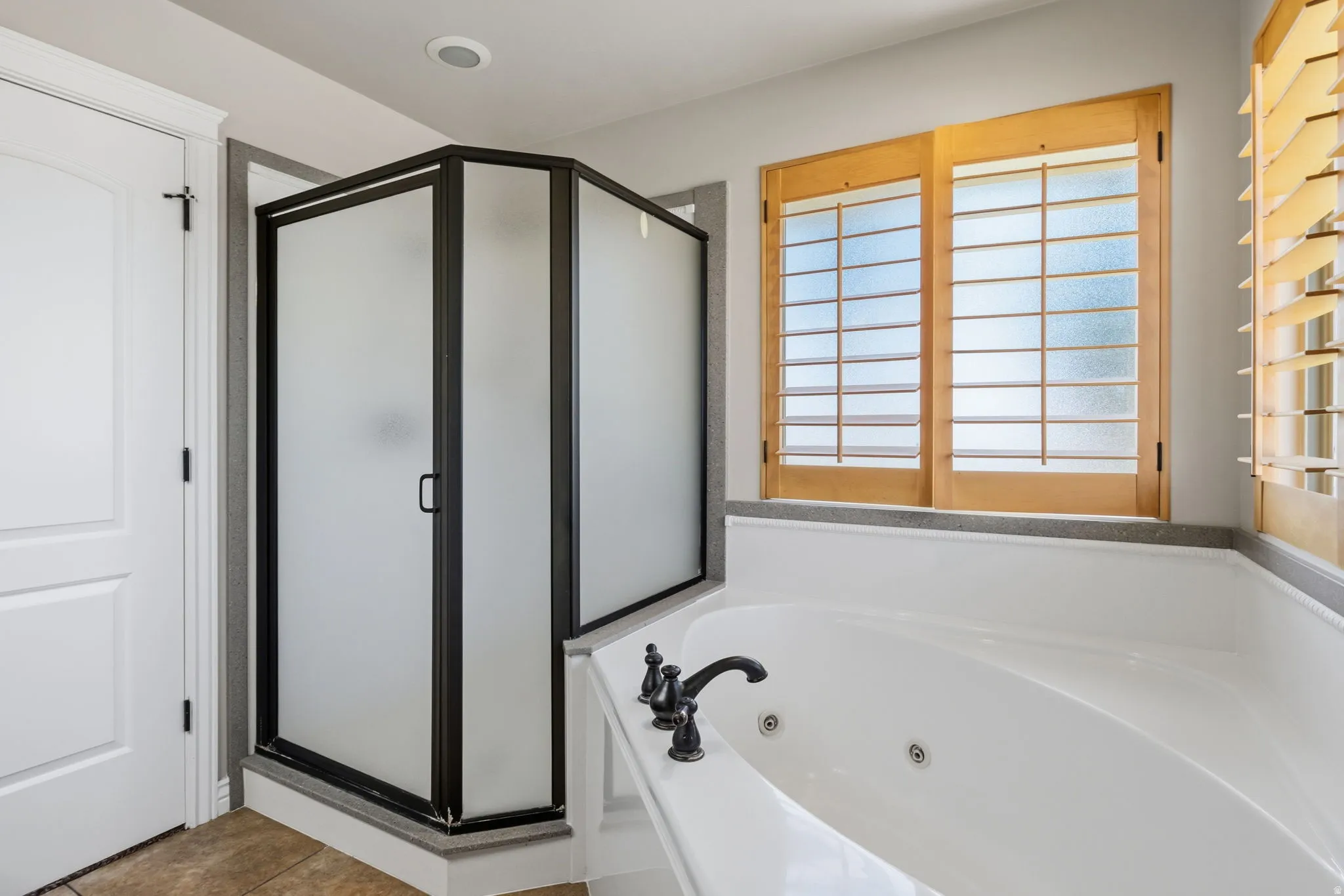 Built-in jetted tub with dark bronze fixtures, frosted glass shower enclosure with dark bronze framing, light wood-tone window shutters, and recessed ceiling lighting