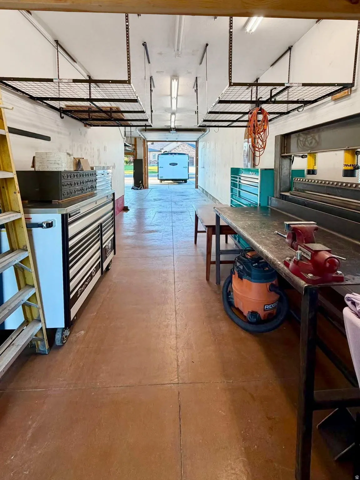 Workshop space featuring a durable concrete floor, overhead storage racks, fluorescent lighting, and a substantial workbench with integrated vises