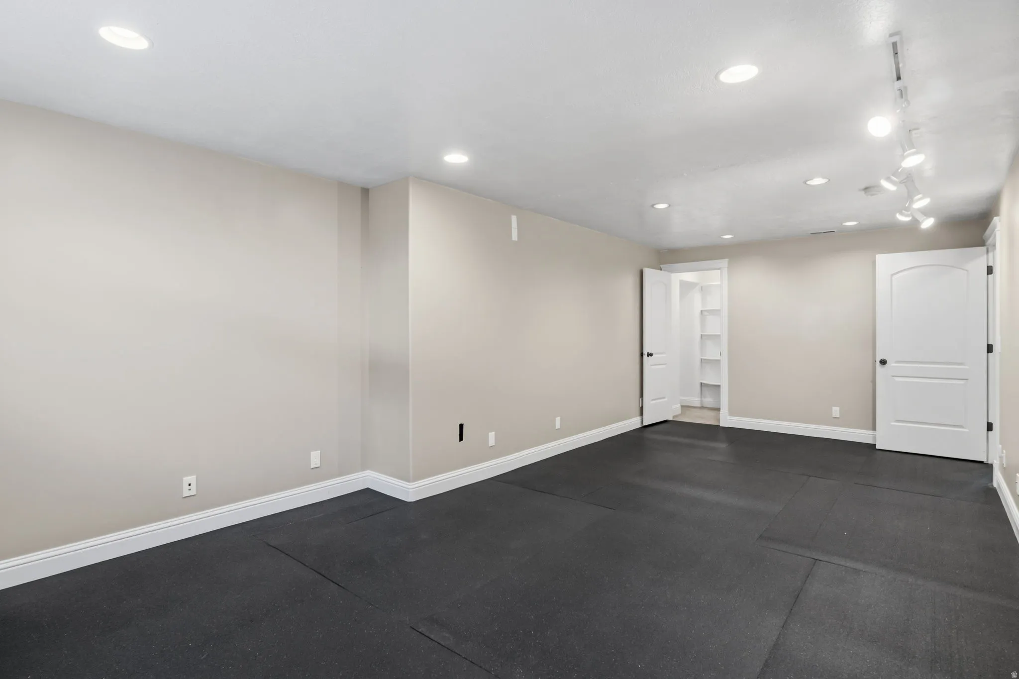 Spacious room featuring dark rubberized flooring, recessed lighting, track lighting, light beige walls, and white trim