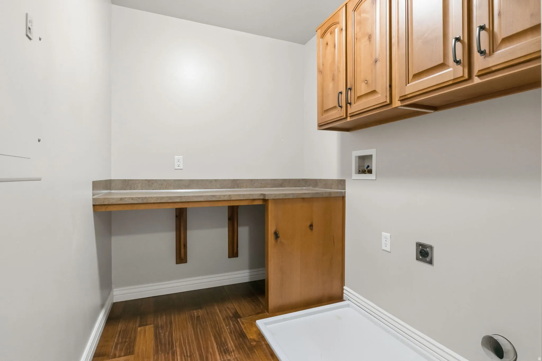 Laundry area featuring wood-finish flooring, light-colored walls, built-in countertop, and wood cabinetry with dark hardware