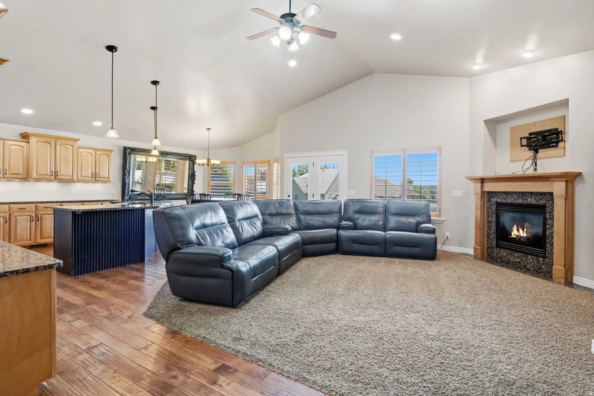 Open-concept living area with vaulted ceilings, a central fireplace featuring a stone surround and wood mantel, wood-finish flooring, and recessed lighting