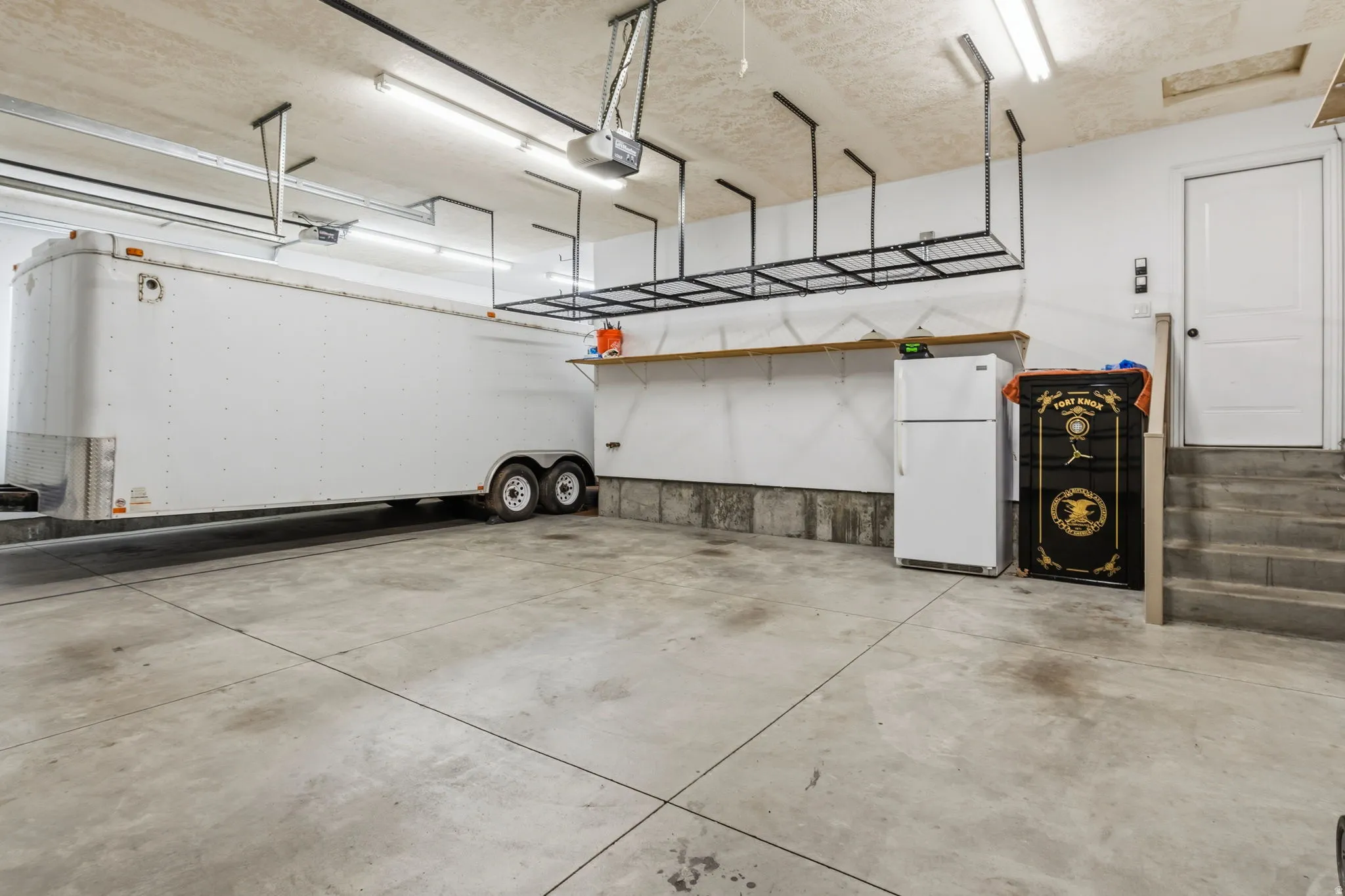 Spacious garage featuring a concrete floor, built-in overhead storage racks, a wooden wall-mounted shelf, fluorescent lighting, and interior access via a white door and concrete steps