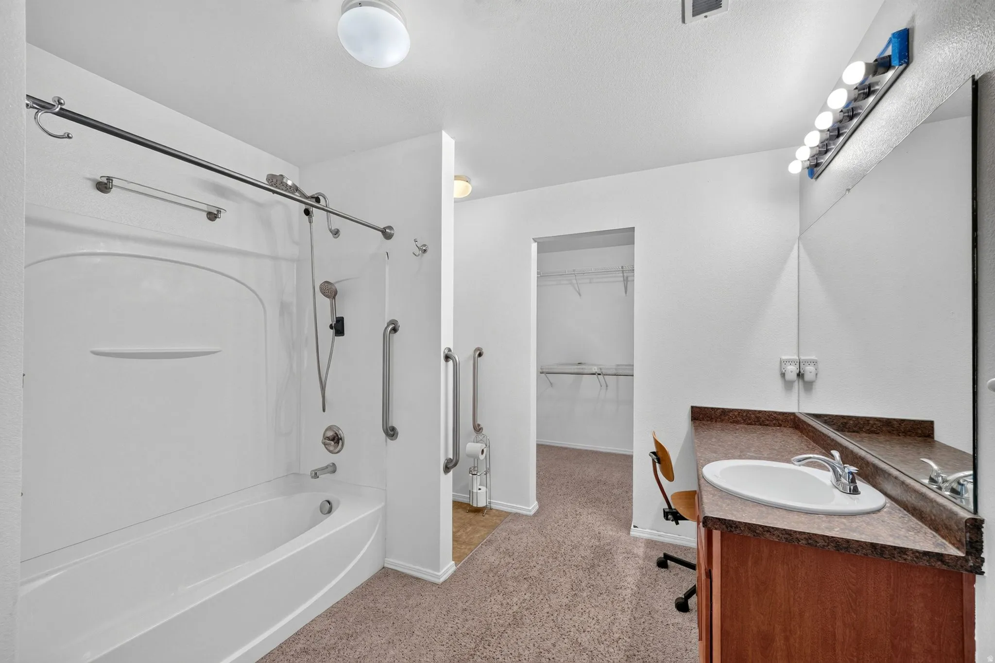 Bathroom featuring a white tub/shower combination with grab bars, a single vanity with an integrated sink and brown countertop, a large wall-mounted mirror, and overhead vanity lighting