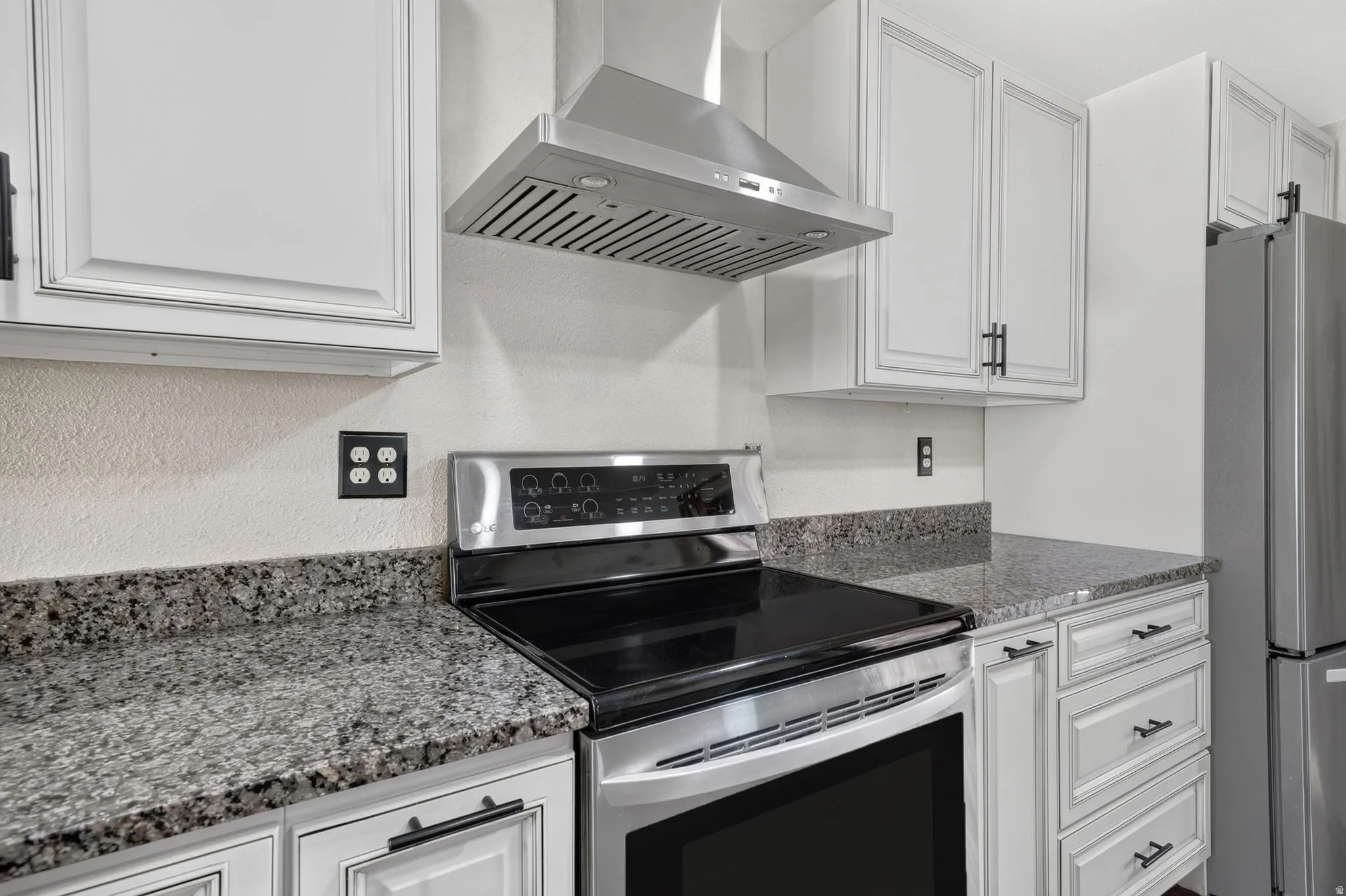 Kitchen featuring stainless steel appliances, a range hood, light cabinetry, dark hardware, and speckled countertops