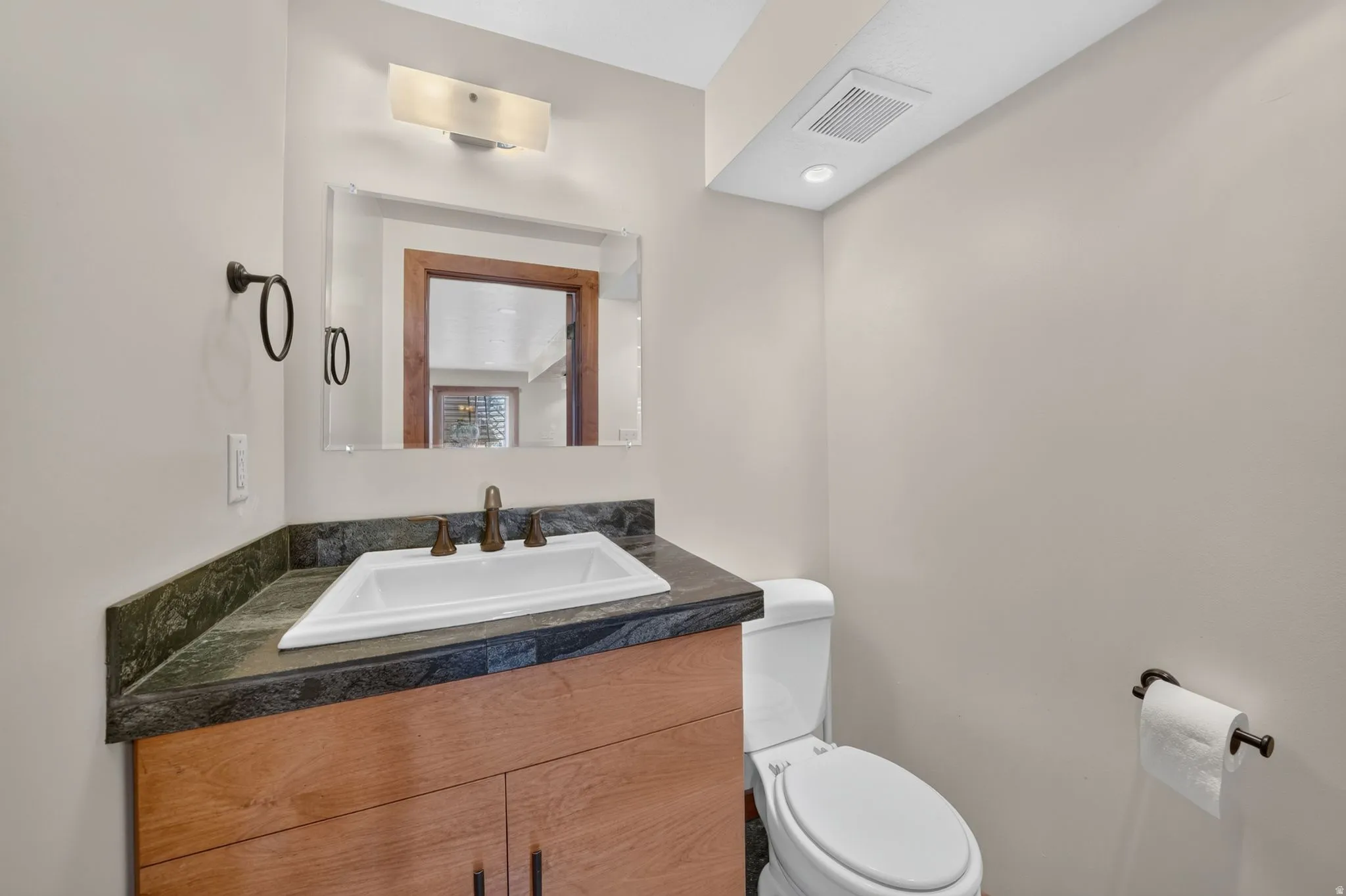 Powder room featuring a wood-finish vanity with a built-in sink, dark stone countertop, wall-mounted mirror, and vanity light fixture