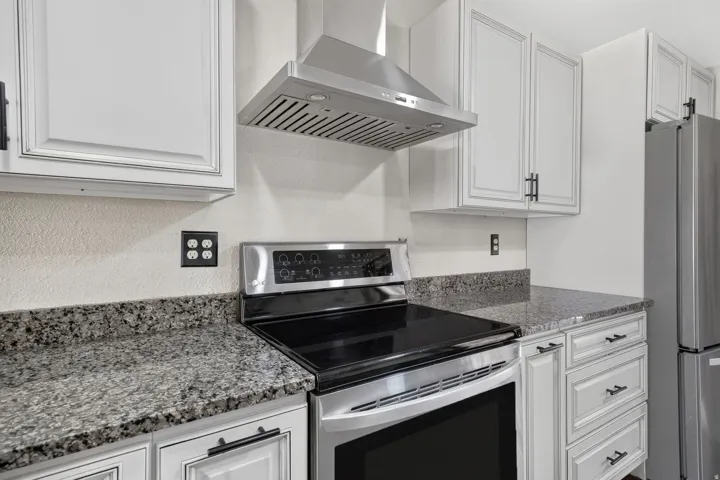 Kitchen featuring stainless steel appliances, a range hood, light cabinetry, dark hardware, and speckled countertops