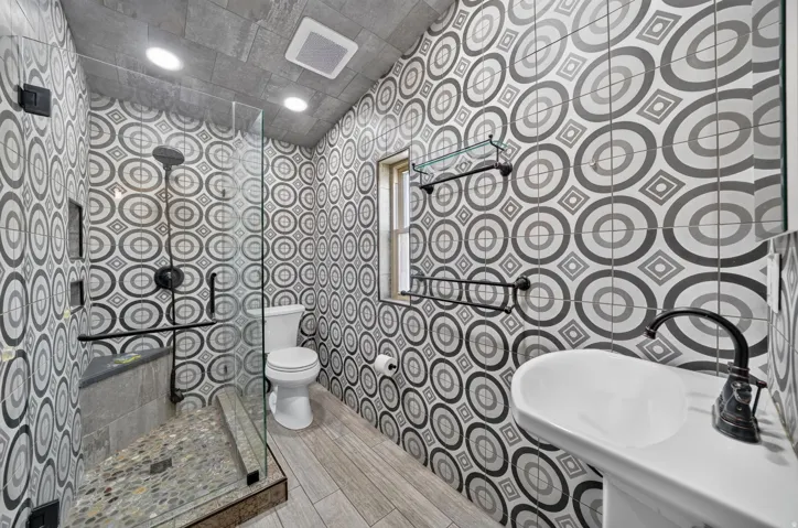 Bathroom featuring patterned wall tiling, a glass-enclosed shower with pebble flooring, a wall-mounted sink with a dark bronze faucet, and wood-finish floor tiles