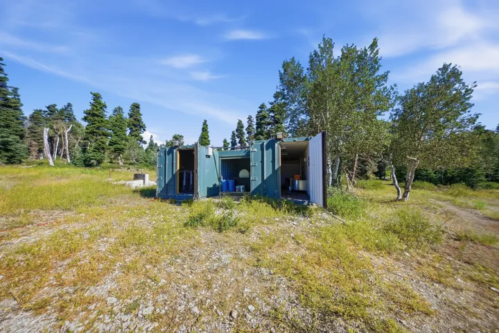 Green storage container structure with open doors, situated on a grassy and rocky terrain