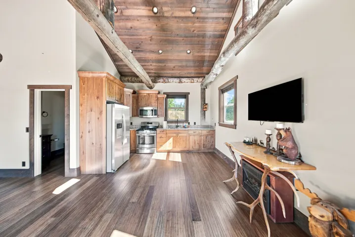 Vaulted wood-plank ceilings with exposed timber beams, wood-finish flooring, and a kitchen featuring wood cabinetry, stainless steel appliances, and a window