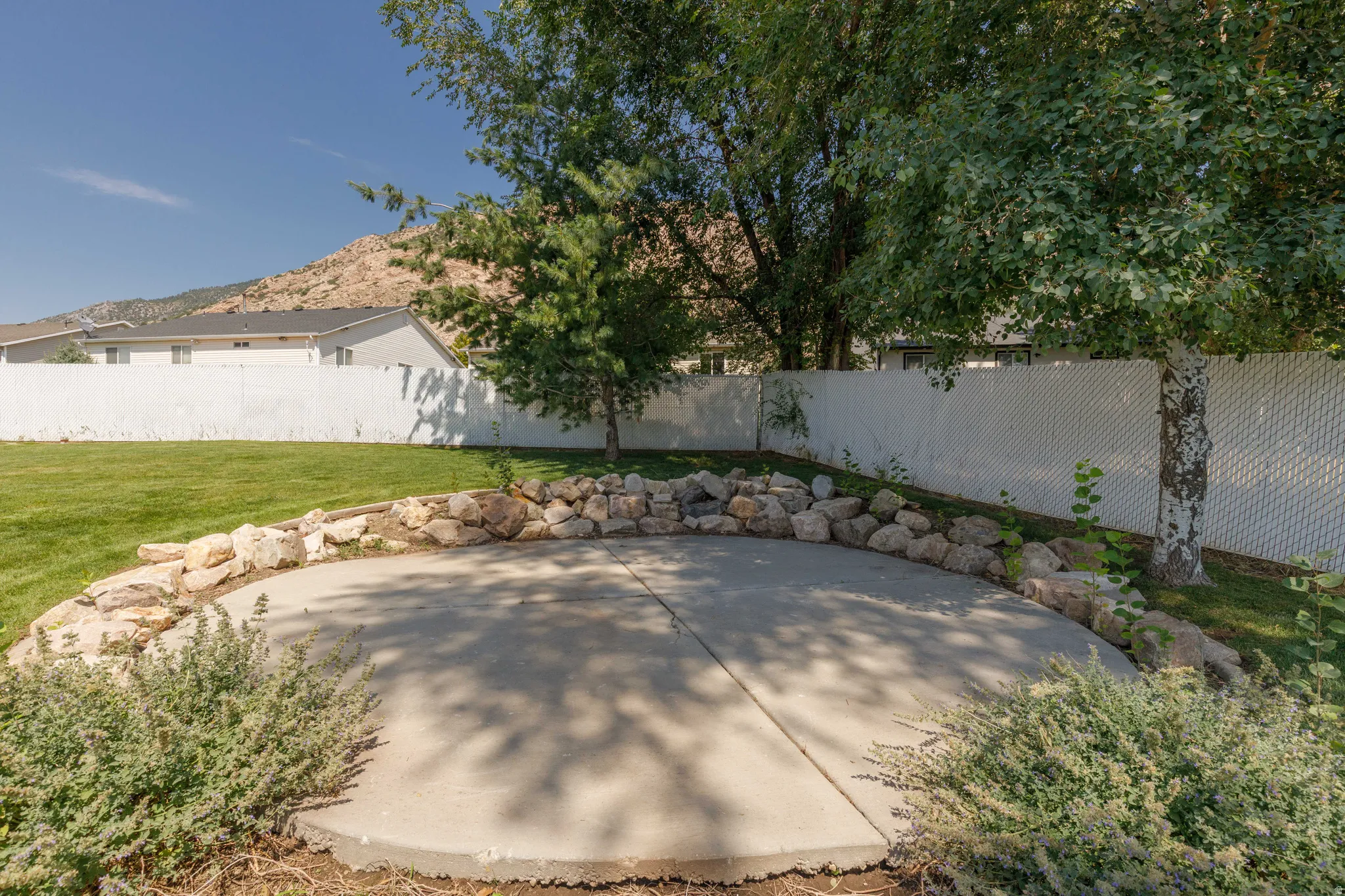 Circular concrete patio surrounded by natural rock landscaping, mature trees, and a spacious green lawn, all enclosed by a white privacy fence