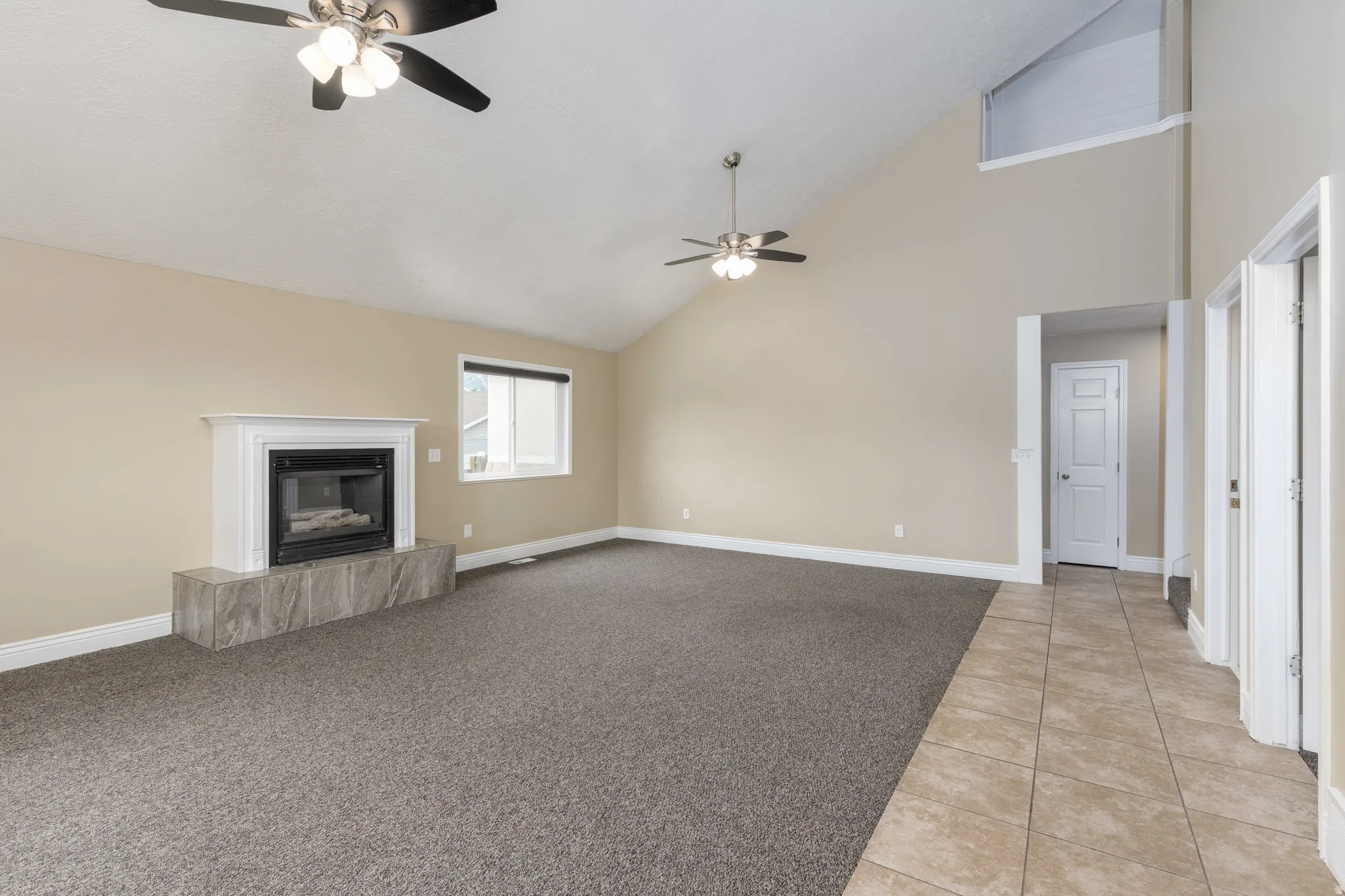 Spacious room featuring vaulted ceilings, a fireplace with a white mantle and a tiled hearth, two ceiling fans, and a combination of carpet and tile flooring
