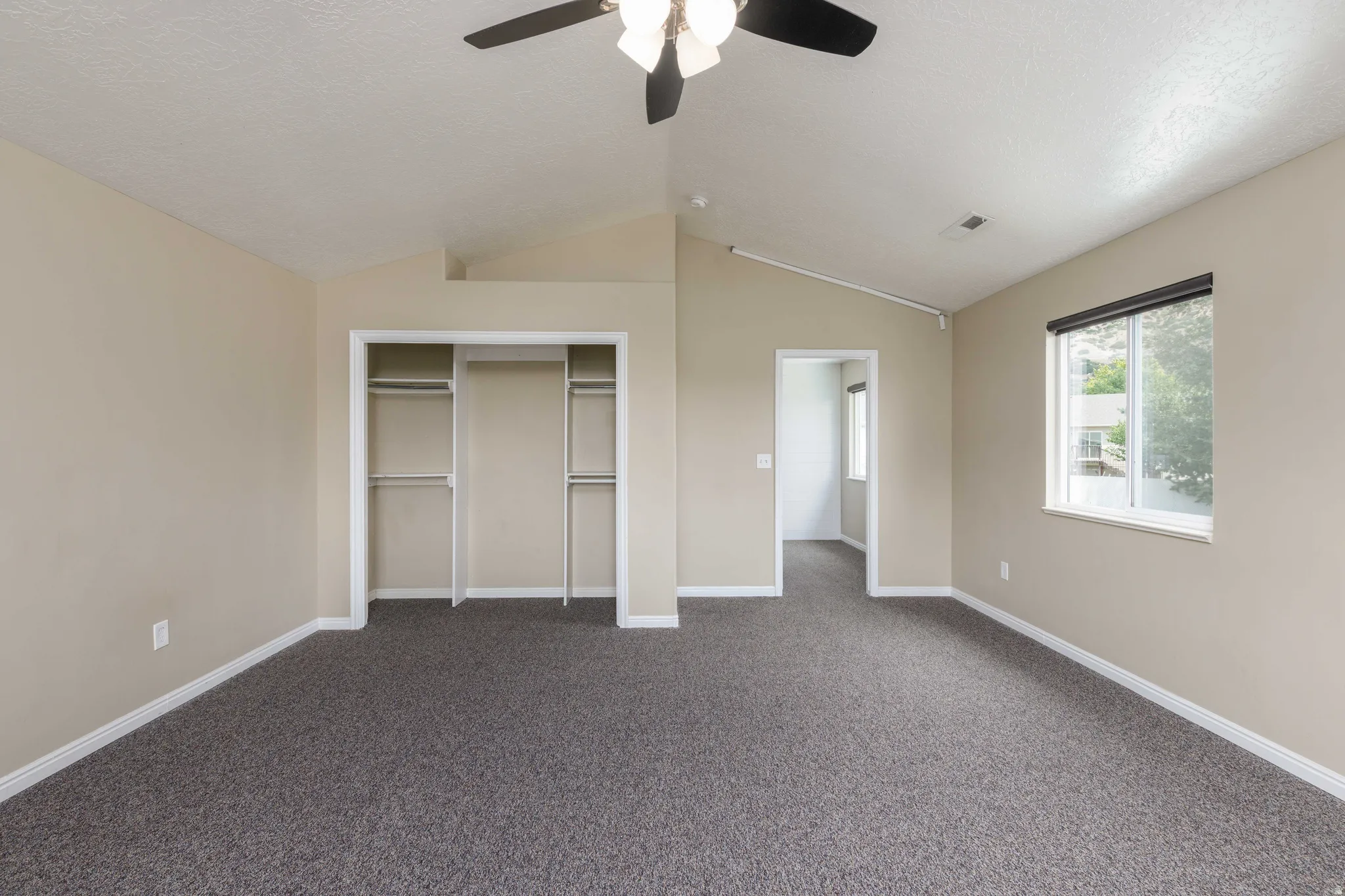 Spacious room featuring vaulted ceilings, a ceiling fan with light fixture, carpeted flooring, a double closet with shelving and hanging rods, and a window with a roller shade