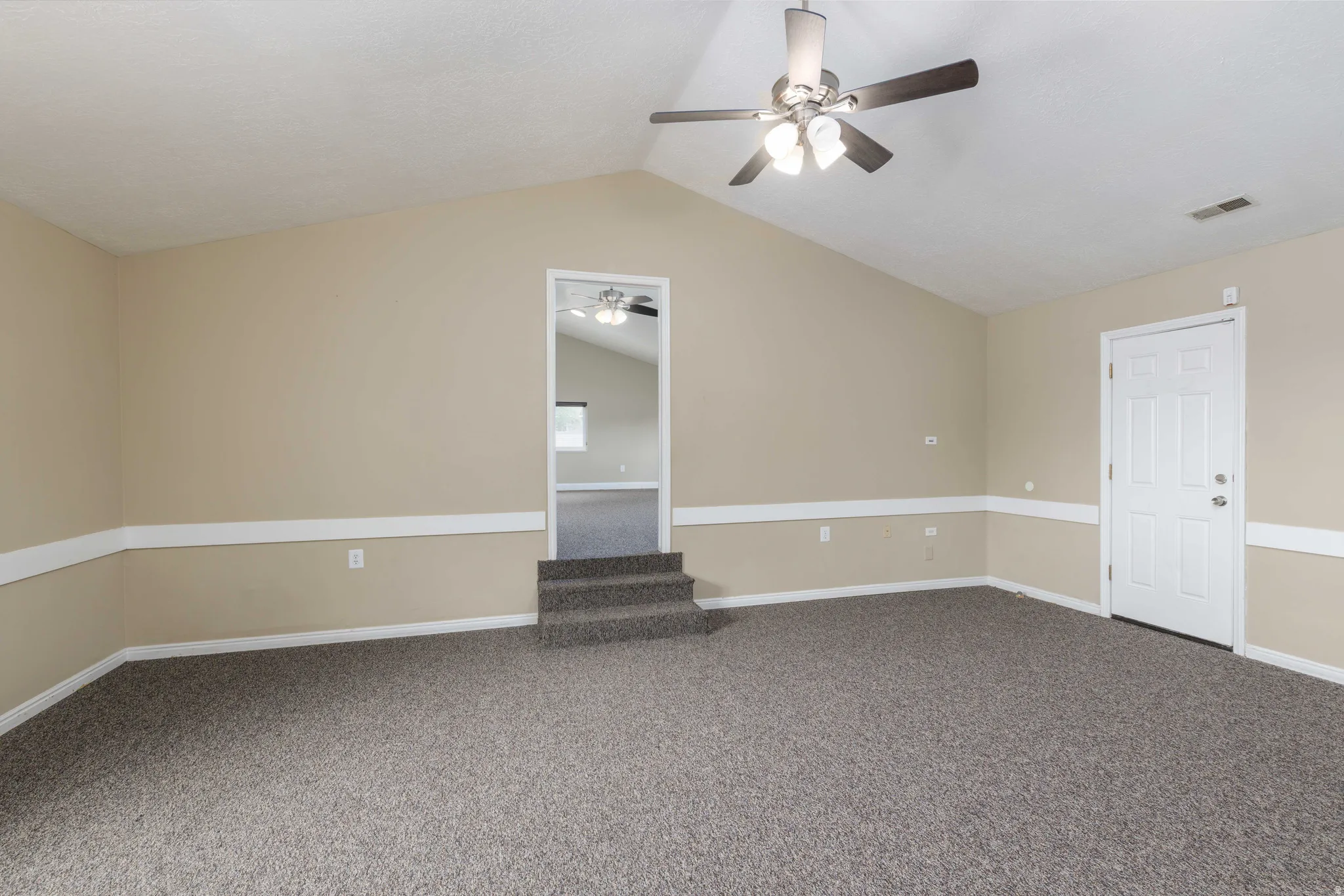 Spacious room featuring vaulted ceilings, textured carpeting, a multi-blade ceiling fan with integrated lighting, and a paneled entry door