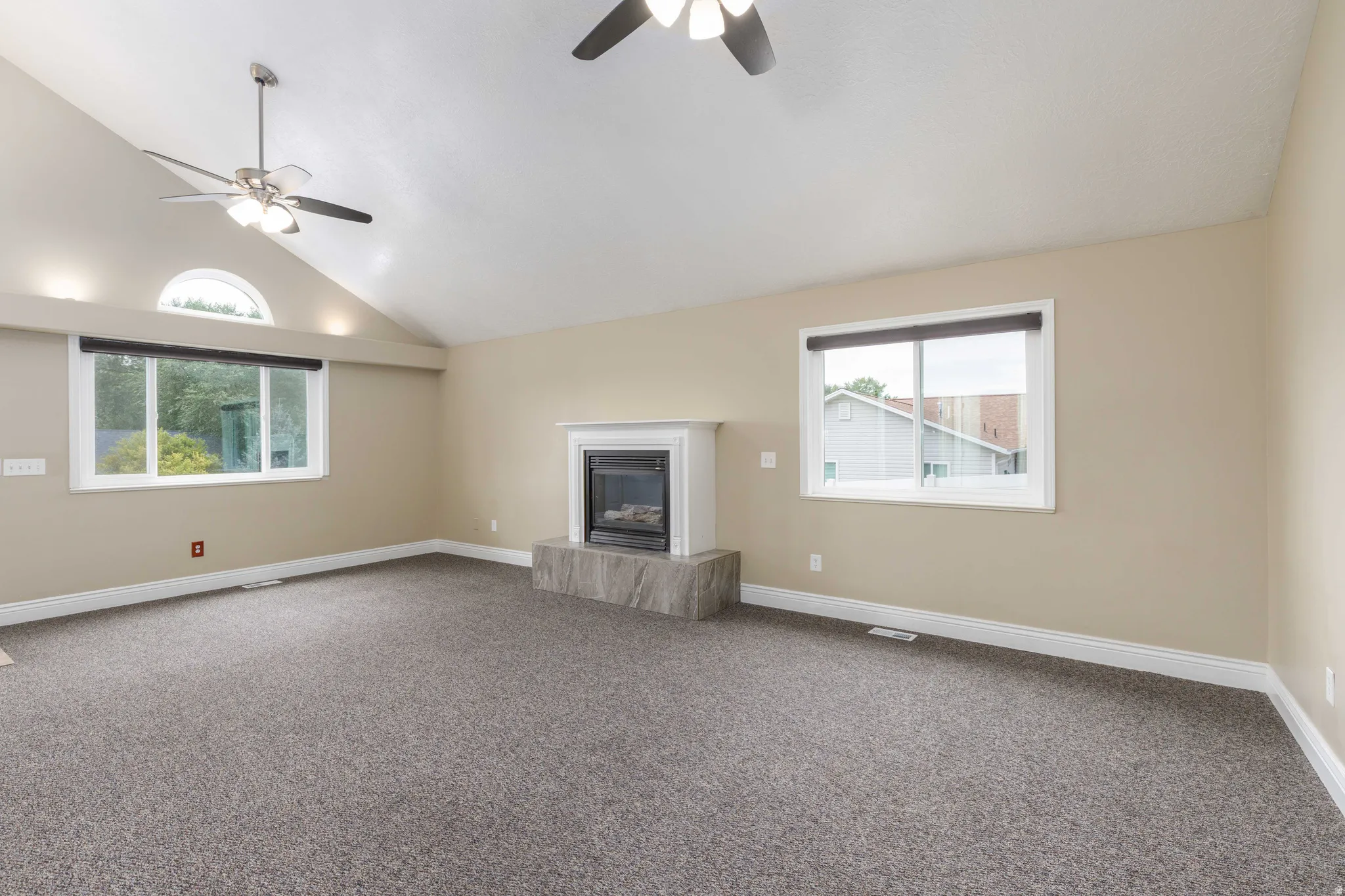 Living area featuring a vaulted ceiling with two ceiling fans, a fireplace with a white mantel and stone-finish hearth, and multiple windows