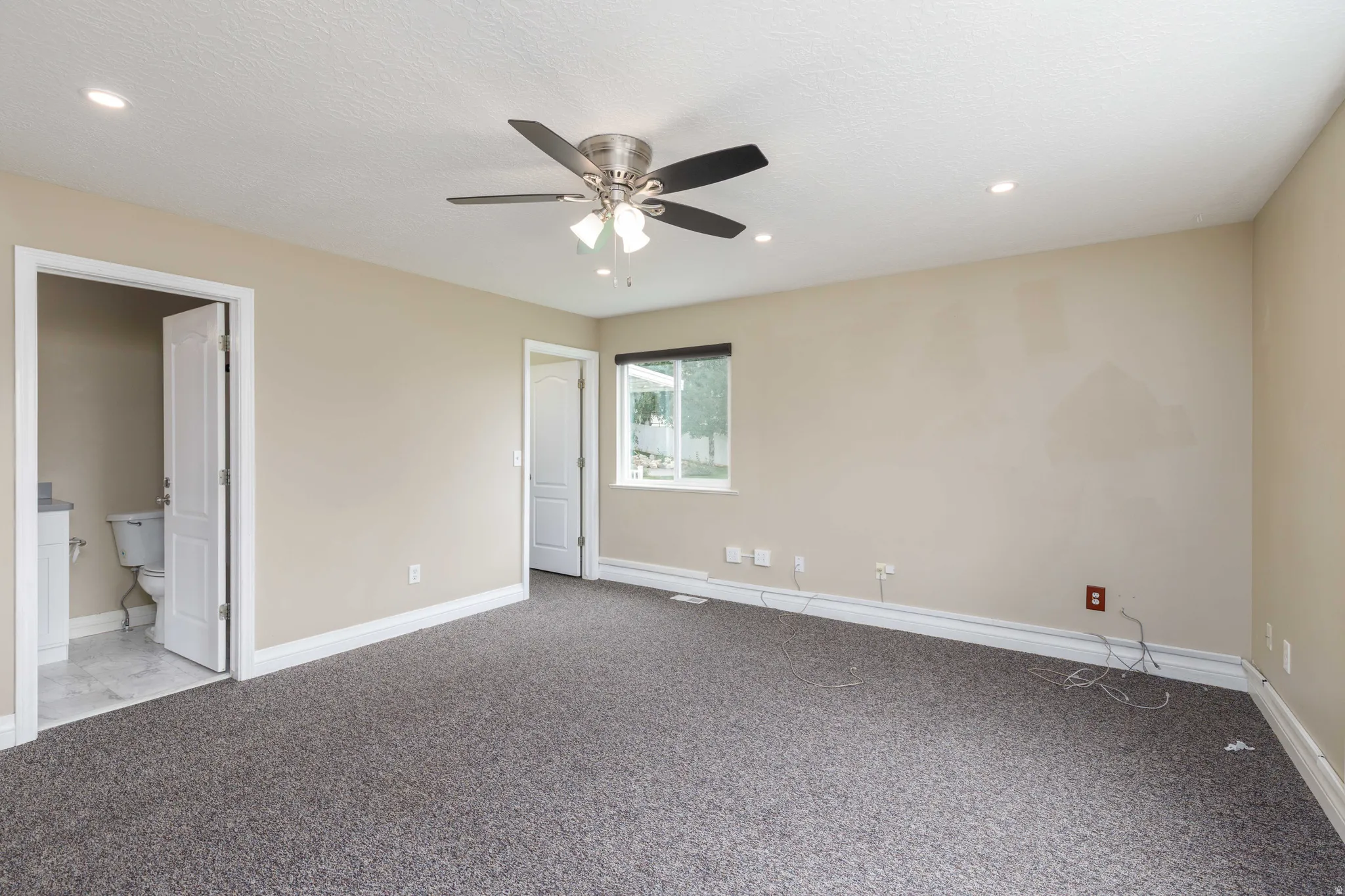 Spacious room featuring neutral wall tones, gray carpeting, a ceiling fan with integrated lighting, recessed lighting, and a window with outdoor landscaping visible