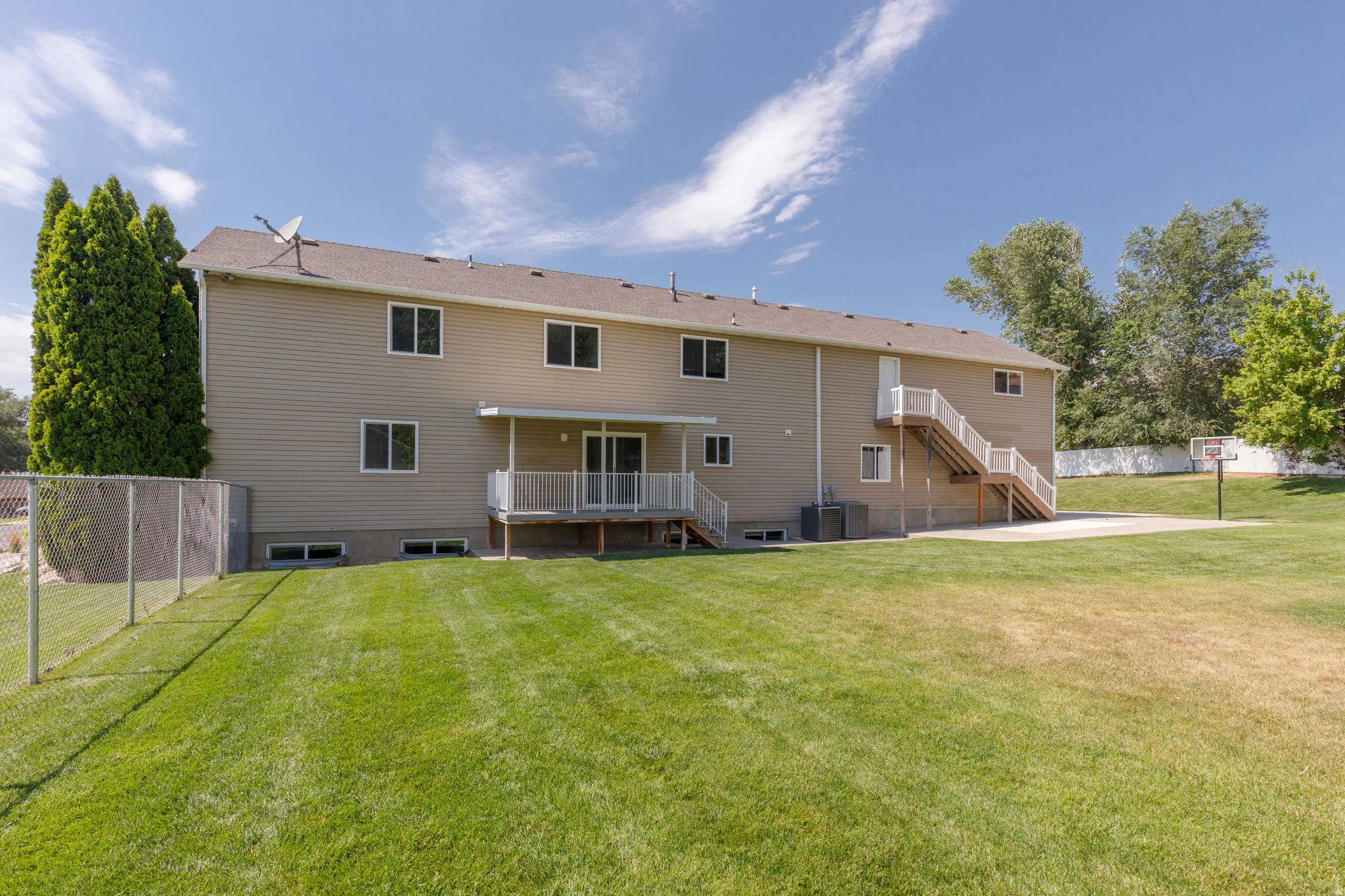 Rear exterior featuring tan siding, a ground-level deck with white railings, a concrete patio, and a chain-link fence