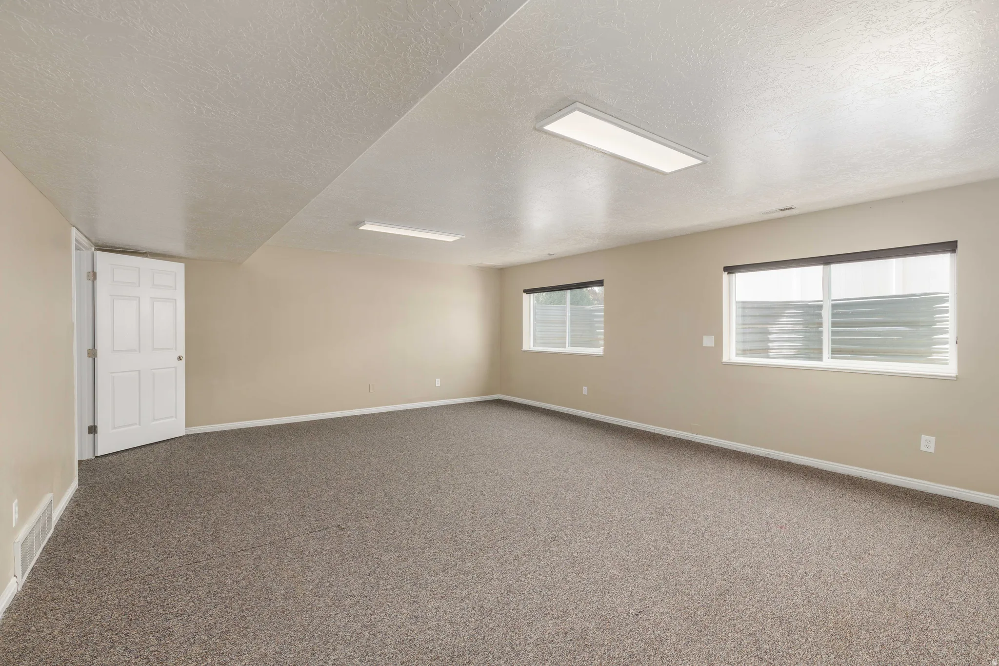 Spacious room featuring neutral carpeting, light beige walls, and two large windows with roller shades