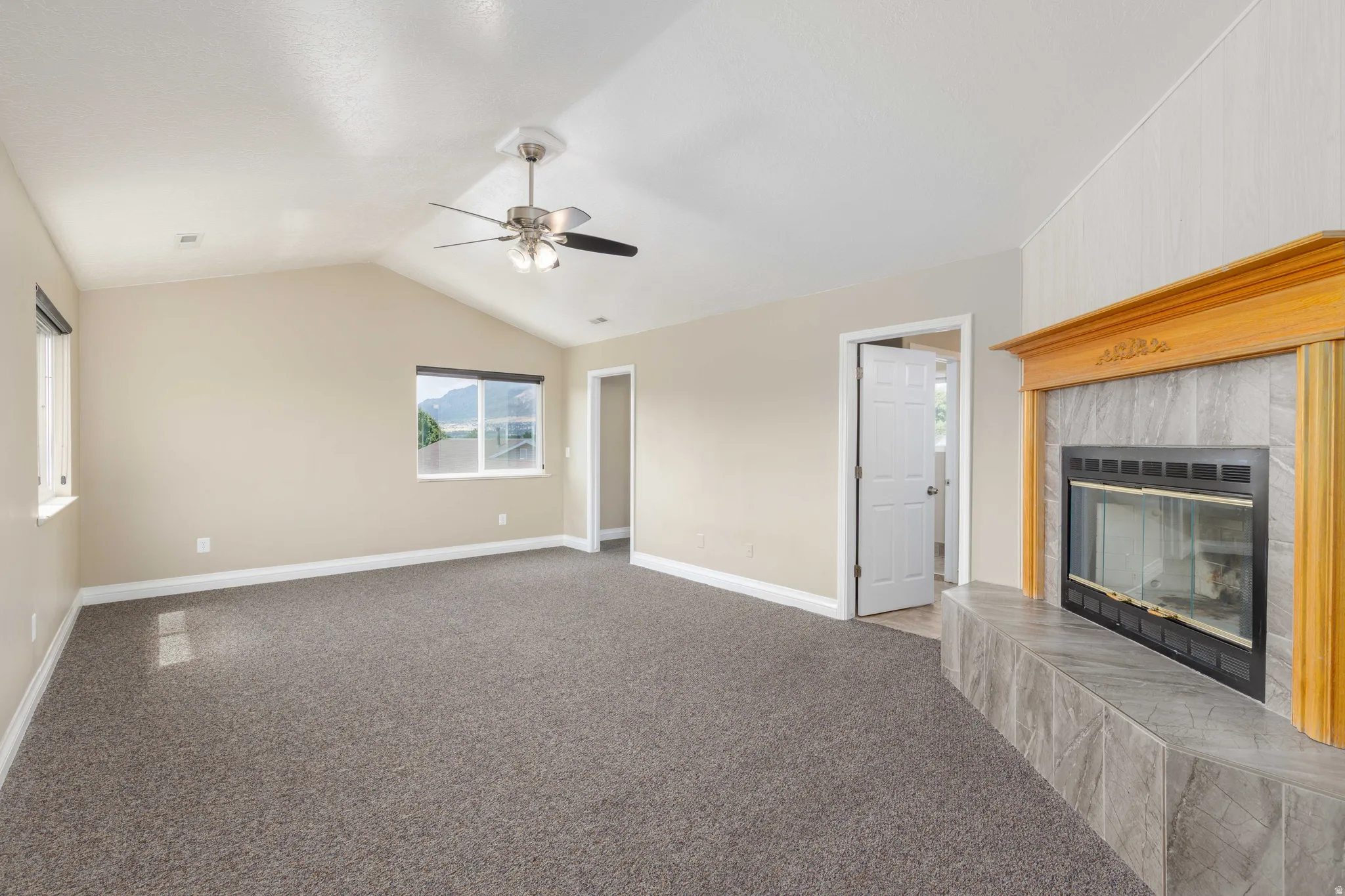 Spacious living area featuring a vaulted ceiling with a ceiling fan, carpet flooring, and a tiled fireplace with a wood mantel