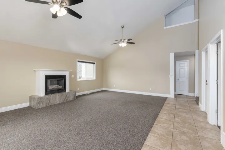 Spacious room featuring vaulted ceilings, a fireplace with a white mantle and a tiled hearth, two ceiling fans, and a combination of carpet and tile flooring