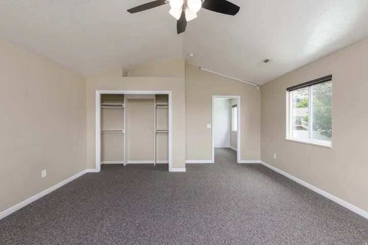Spacious room featuring vaulted ceilings, a ceiling fan with light fixture, carpeted flooring, a double closet with shelving and hanging rods, and a window with a roller shade