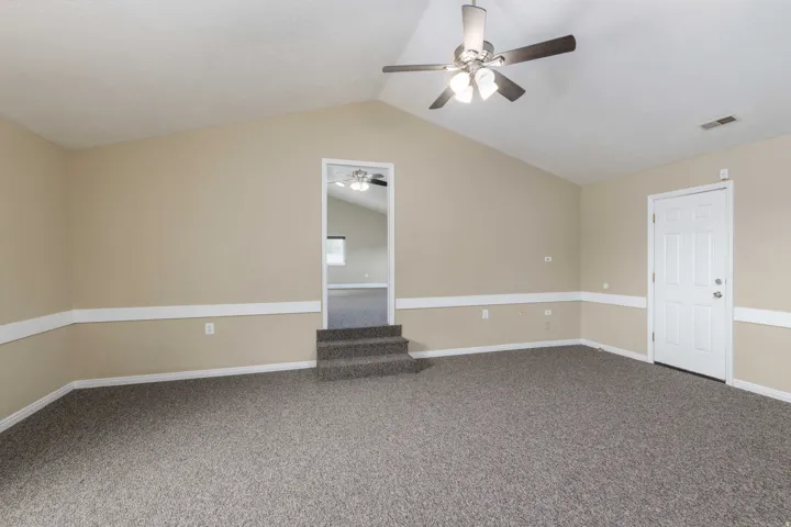 Spacious room featuring vaulted ceilings, textured carpeting, a multi-blade ceiling fan with integrated lighting, and a paneled entry door