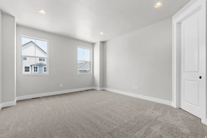 Spacious room featuring neutral carpeting, light gray wall tones, two large windows, recessed ceiling lighting, and a white paneled door with black hardware