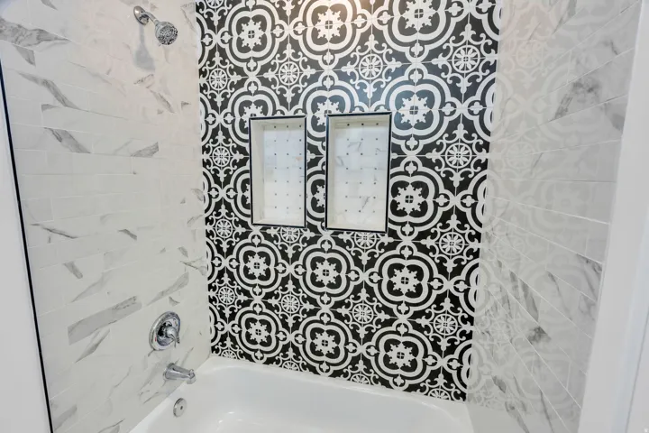 Bathtub and shower combination featuring black and white patterned tile, two recessed shower niches, white subway tile with gray veining, and chrome fixtures