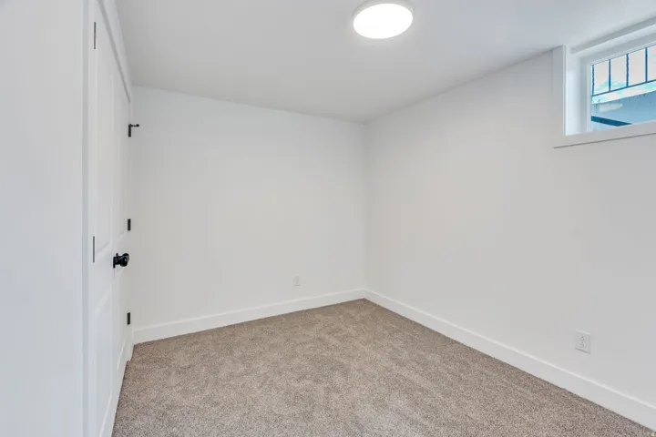 Room featuring neutral carpet flooring, a flush-mount ceiling light, white baseboards, a white door with black hardware, and an egress window