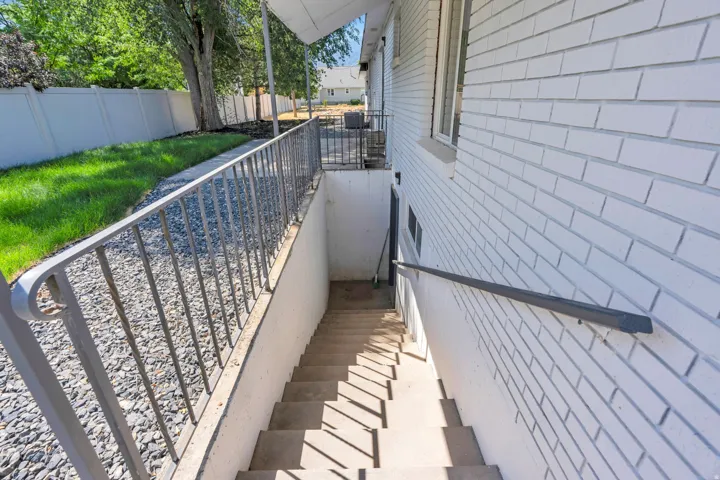 Exterior basement access featuring concrete steps, a metal handrail, and a white brick-style wall