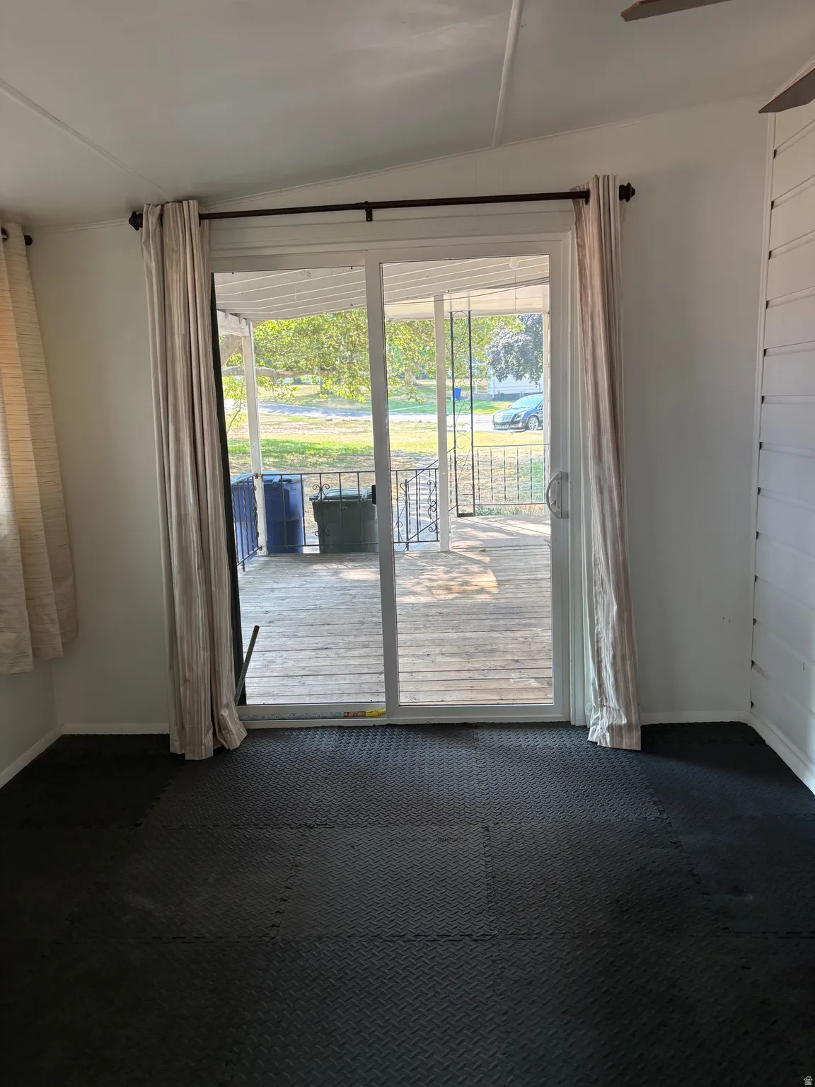 Sliding glass doors open to a spacious deck with wood-finish flooring, framed by neutral curtain panels and complemented by dark carpet flooring in the interior room