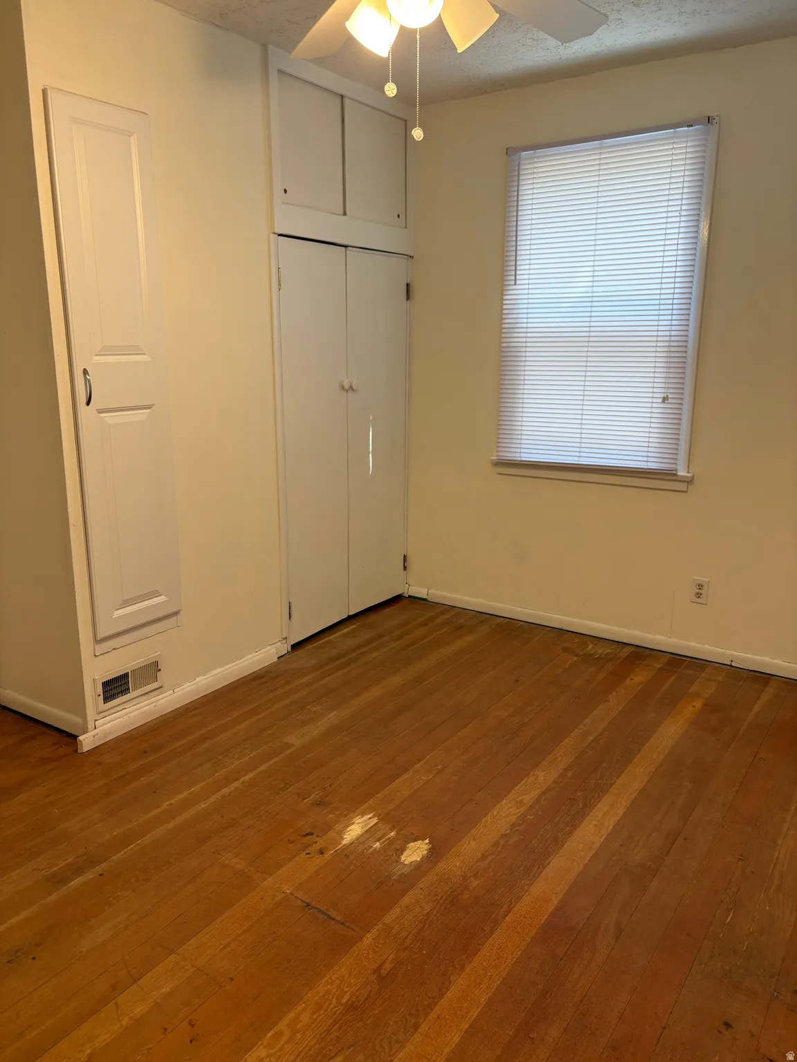 Bright, light-filled bedroom showcasing hardwood flooring, large window with blinds providing natural illumination, overhead ceiling fixture, and clean wall surfaces with electrical outlet