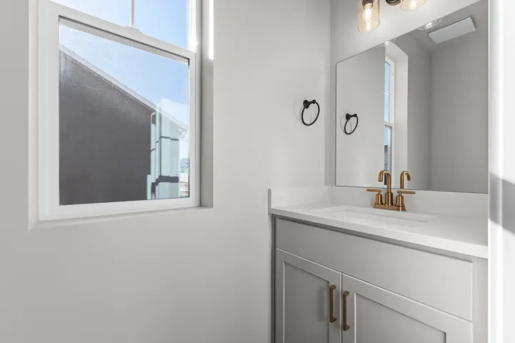 White framed window, light gray vanity with panel-style doors, white countertop, integrated sink, and brass-finish faucet