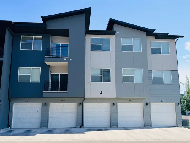 Modern multi-story building featuring a distinct architectural profile, contrasting gray and blue exterior siding, multiple balconies with black metal railings, and individual ground-level garages