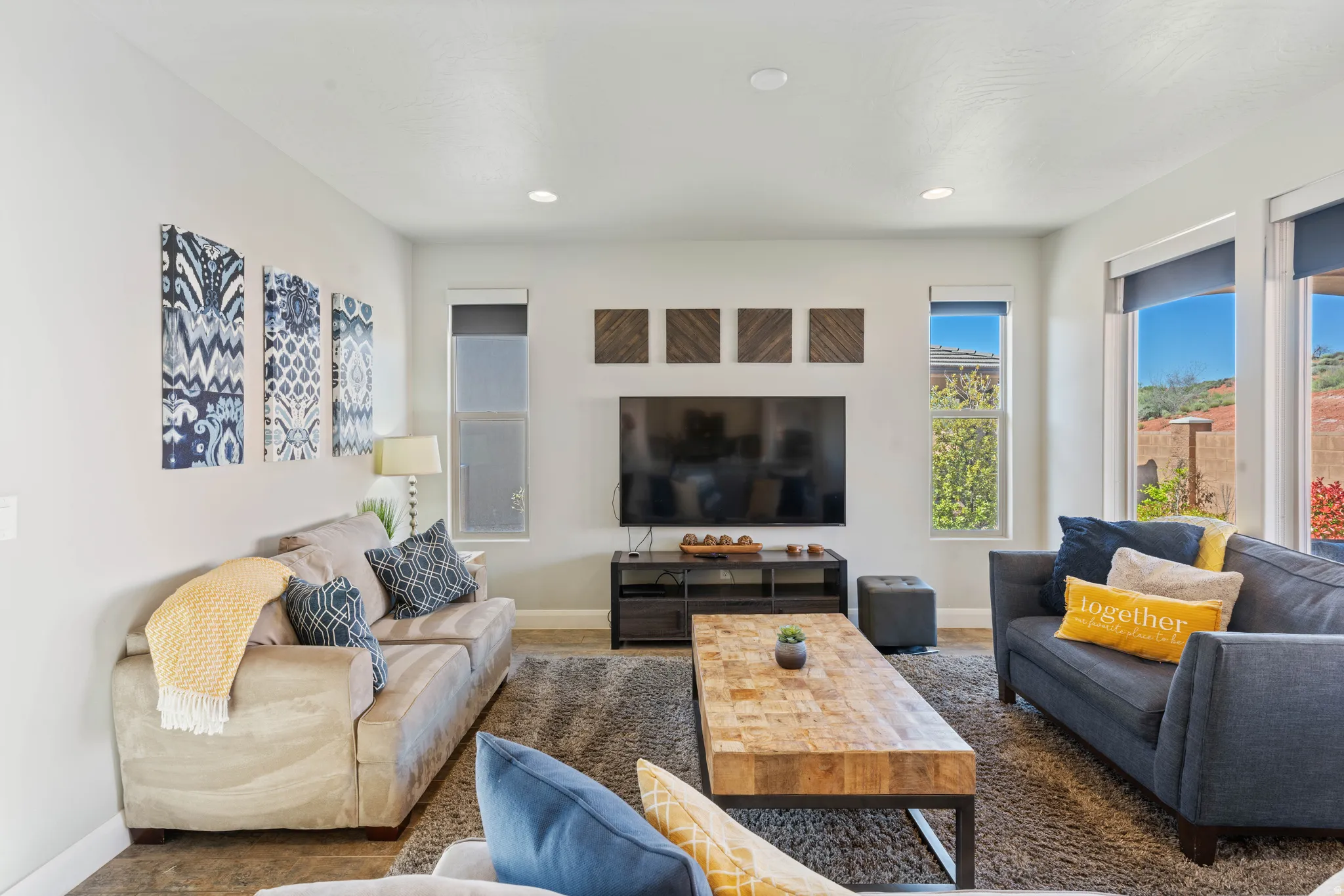 Spacious living area featuring light gray walls, recessed lighting, large windows with roller shades, and dark wood-finish flooring partially covered by a gray area rug