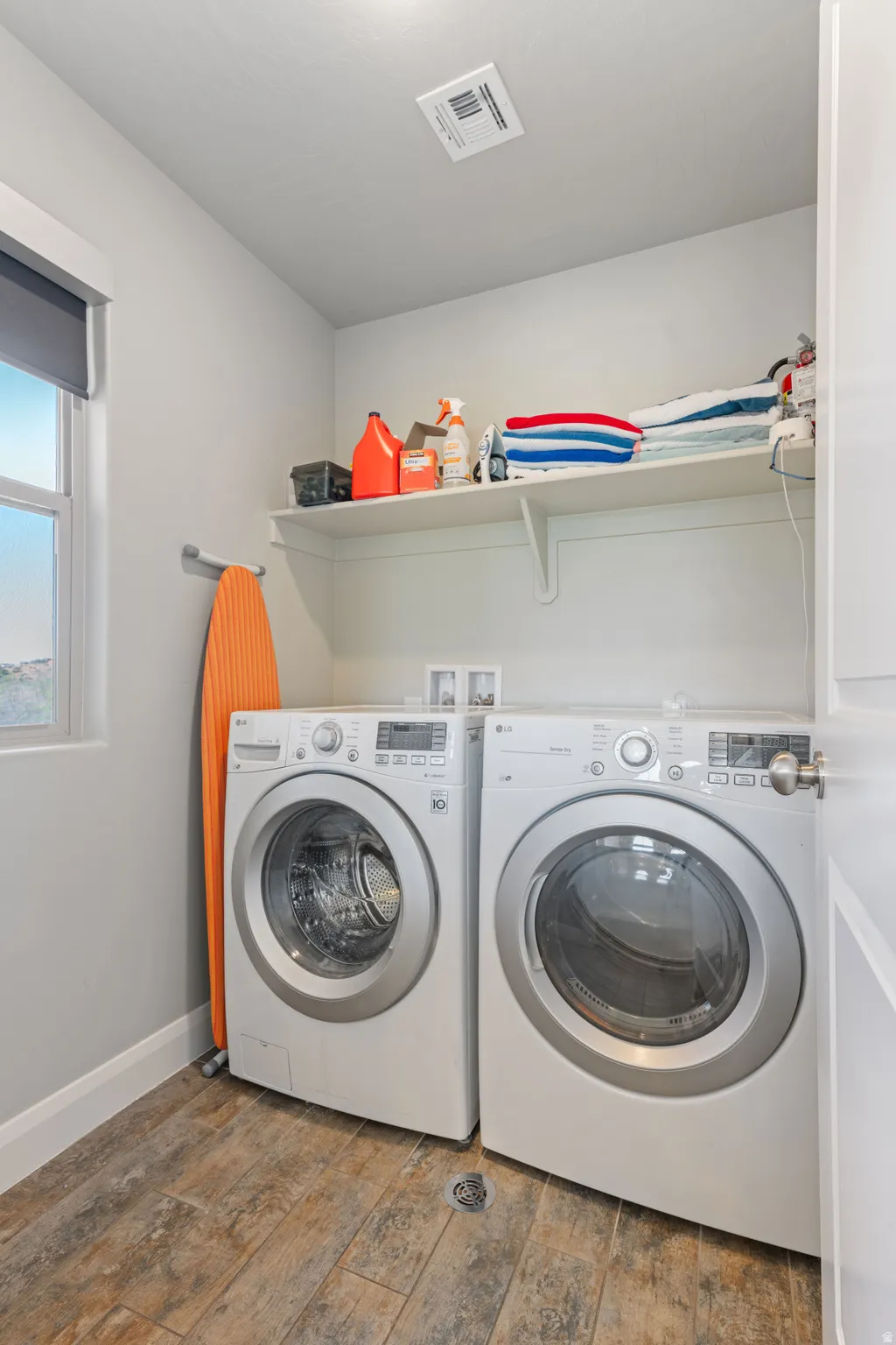 Dedicated laundry area featuring wood-look tile flooring, a window with a roller shade, and an overhead shelf for storage