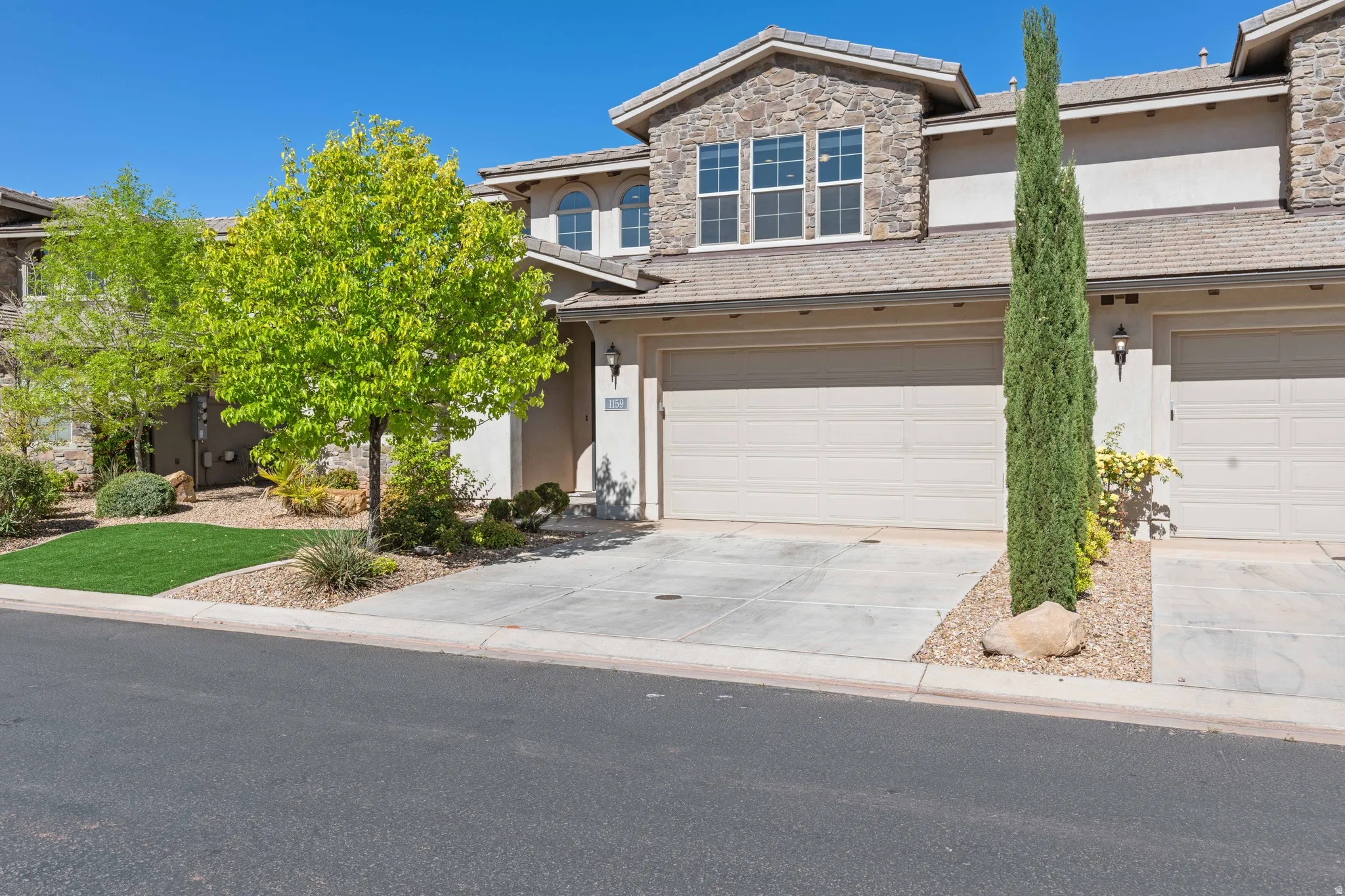 Stone and stucco exterior featuring a two-car garage, concrete driveway, and low-maintenance landscaping with mature trees and shrubs