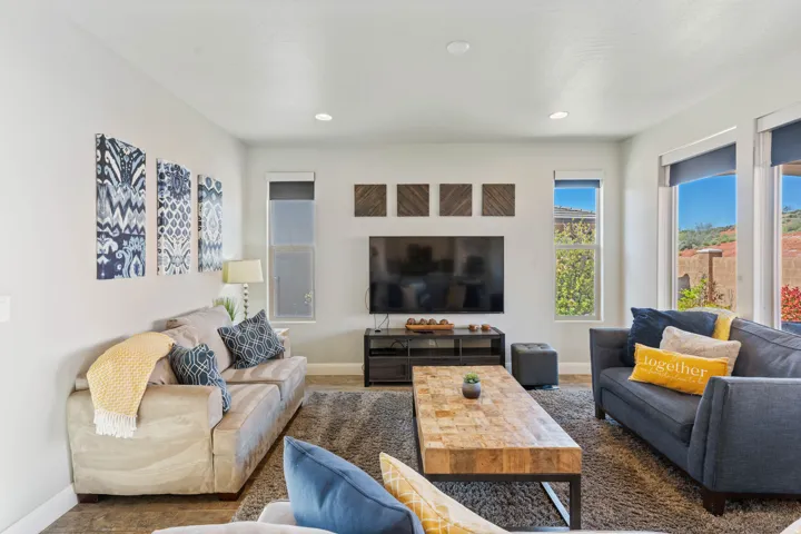 Spacious living area featuring light gray walls, recessed lighting, large windows with roller shades, and dark wood-finish flooring partially covered by a gray area rug