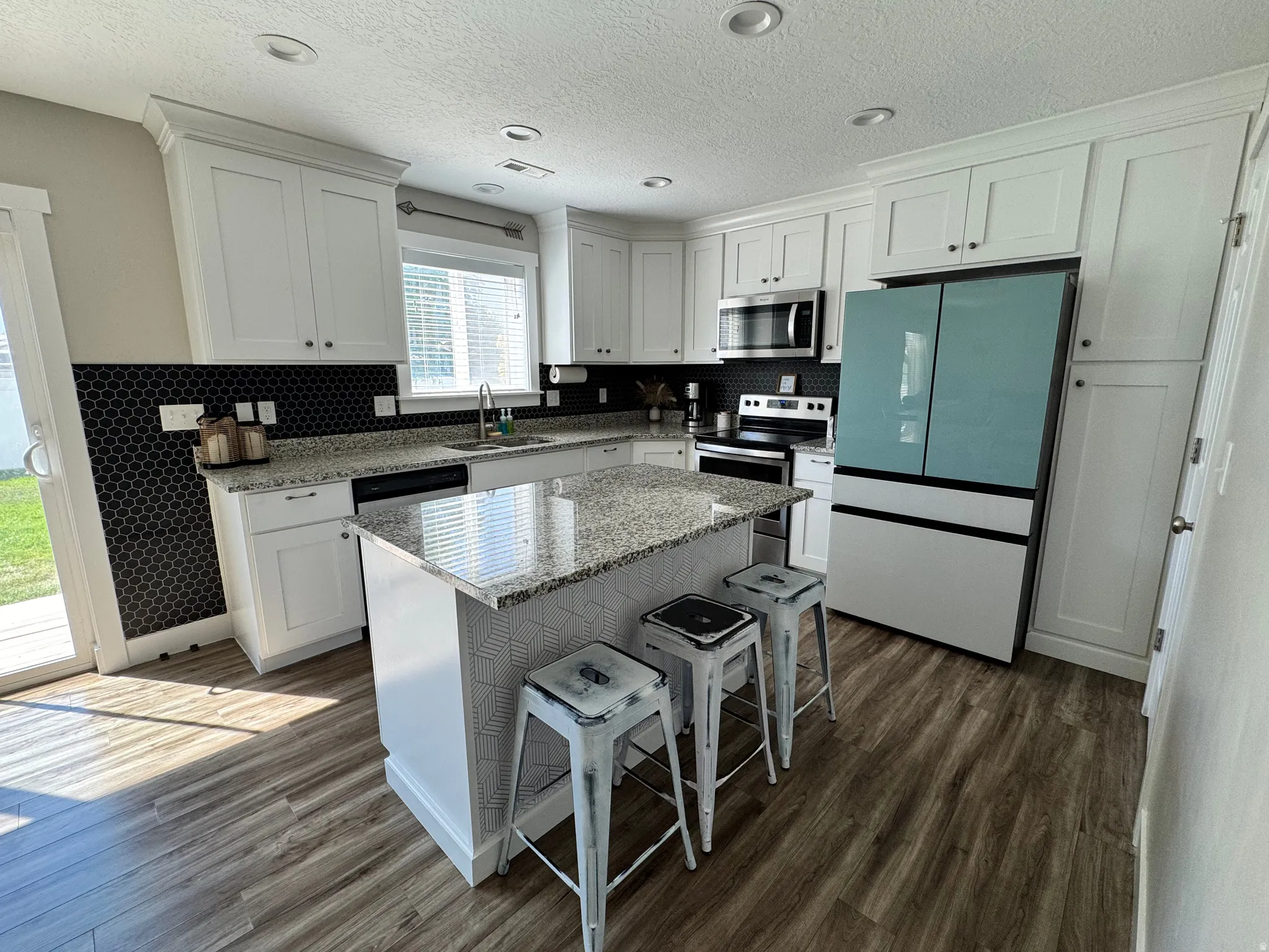 Modern kitchen featuring wood-finish flooring, white cabinetry, a large kitchen island with a stone countertop, a full suite of stainless steel appliances, and a black honeycomb tile backsplash