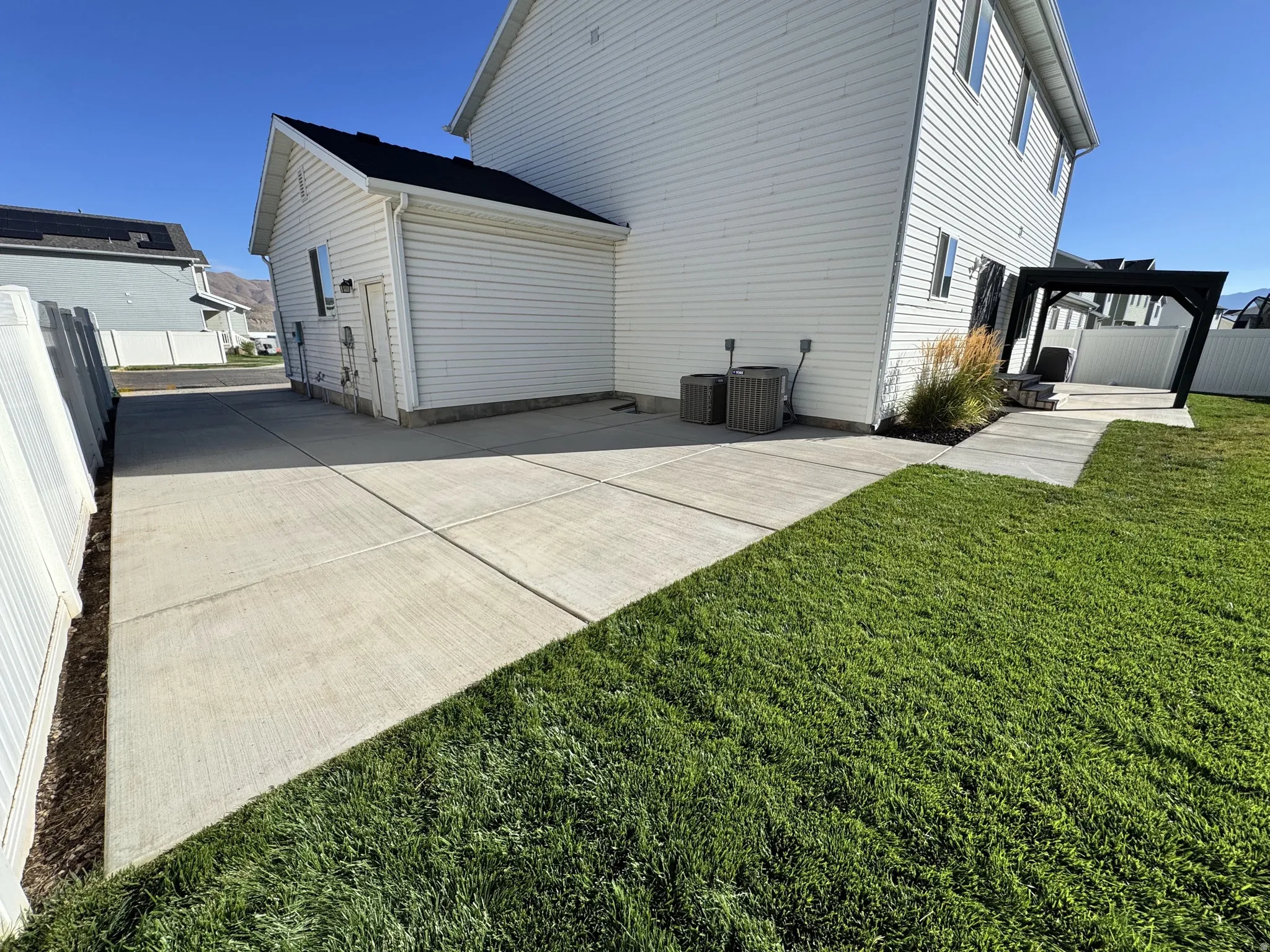 Expansive concrete patio area with a pergola-covered section, featuring a neatly maintained lawn and white privacy fencing