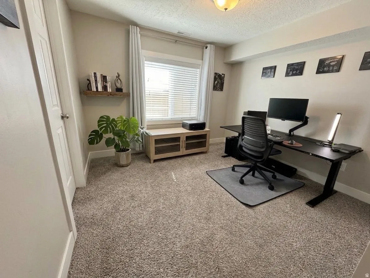Dedicated home office featuring neutral carpet flooring, expansive desk workspace with black ergonomic chair, and abundant natural light from large window with plantation shutters.  AI enhanced design concept.