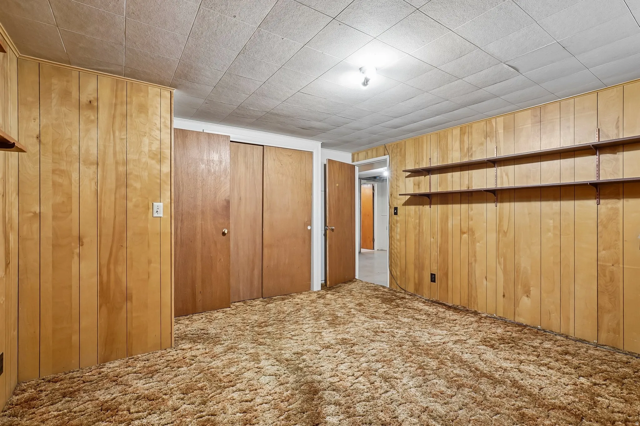 Room featuring wood paneling, shaggy carpet flooring, a ceiling with acoustic tiles, and two sets of bi-fold closet doors