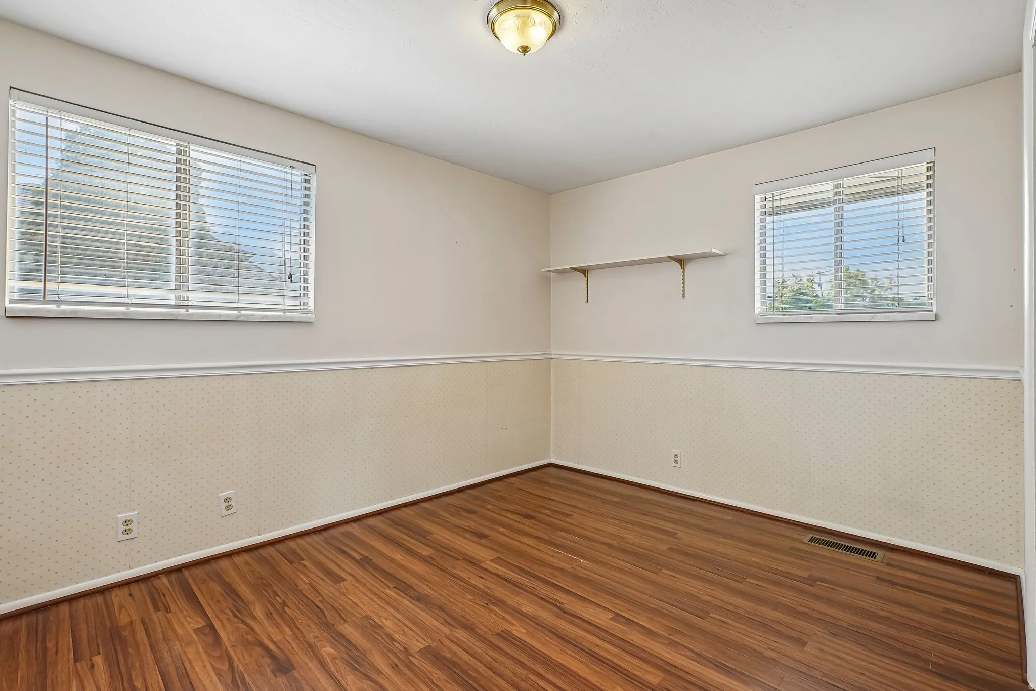 Room with wood-finish flooring, two windows with blinds, a built-in wall shelf, and wainscoting