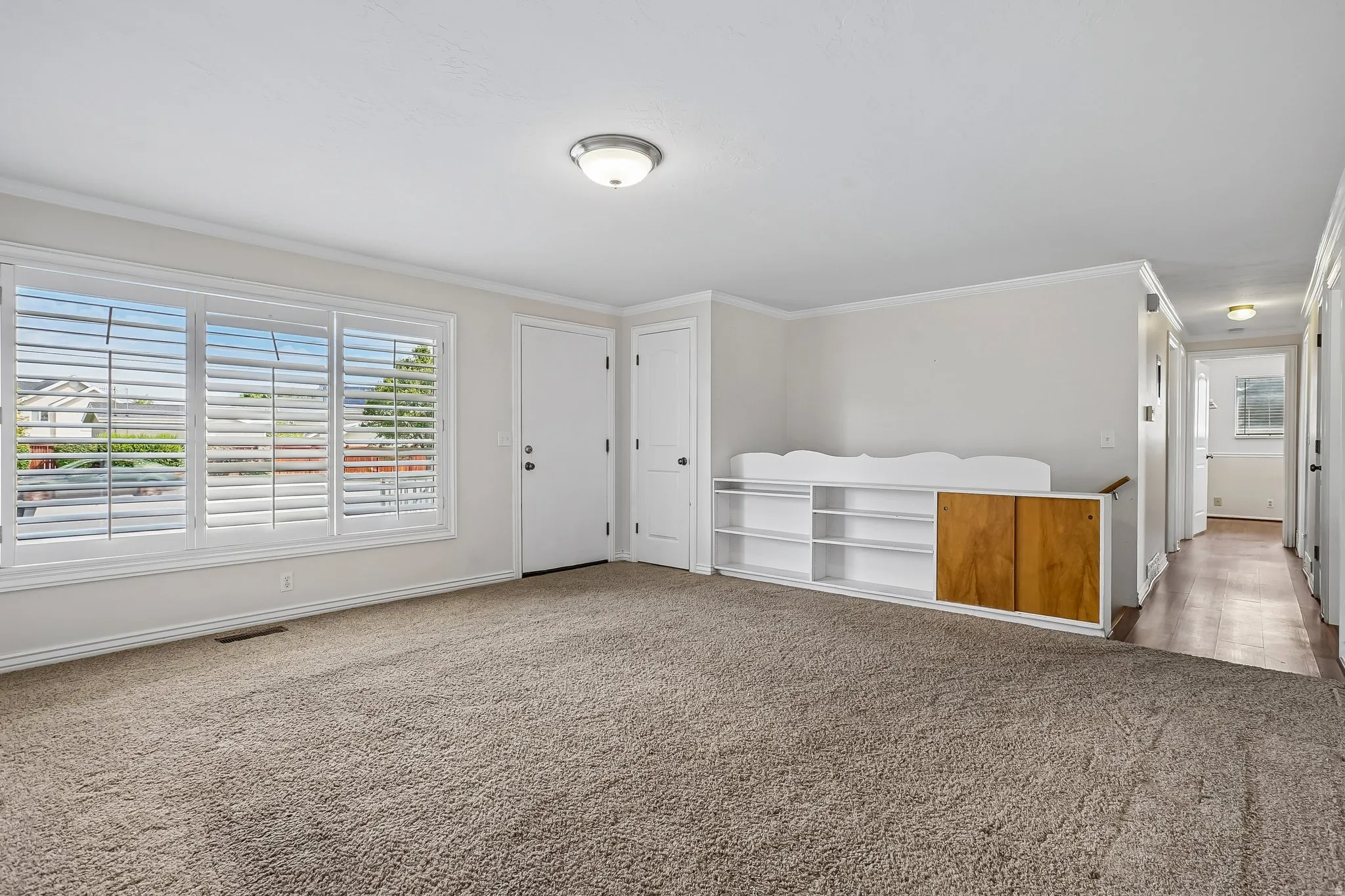 Spacious room featuring neutral carpeting, a large window with white plantation shutters, and a built-in shelving unit with wood-finish doors