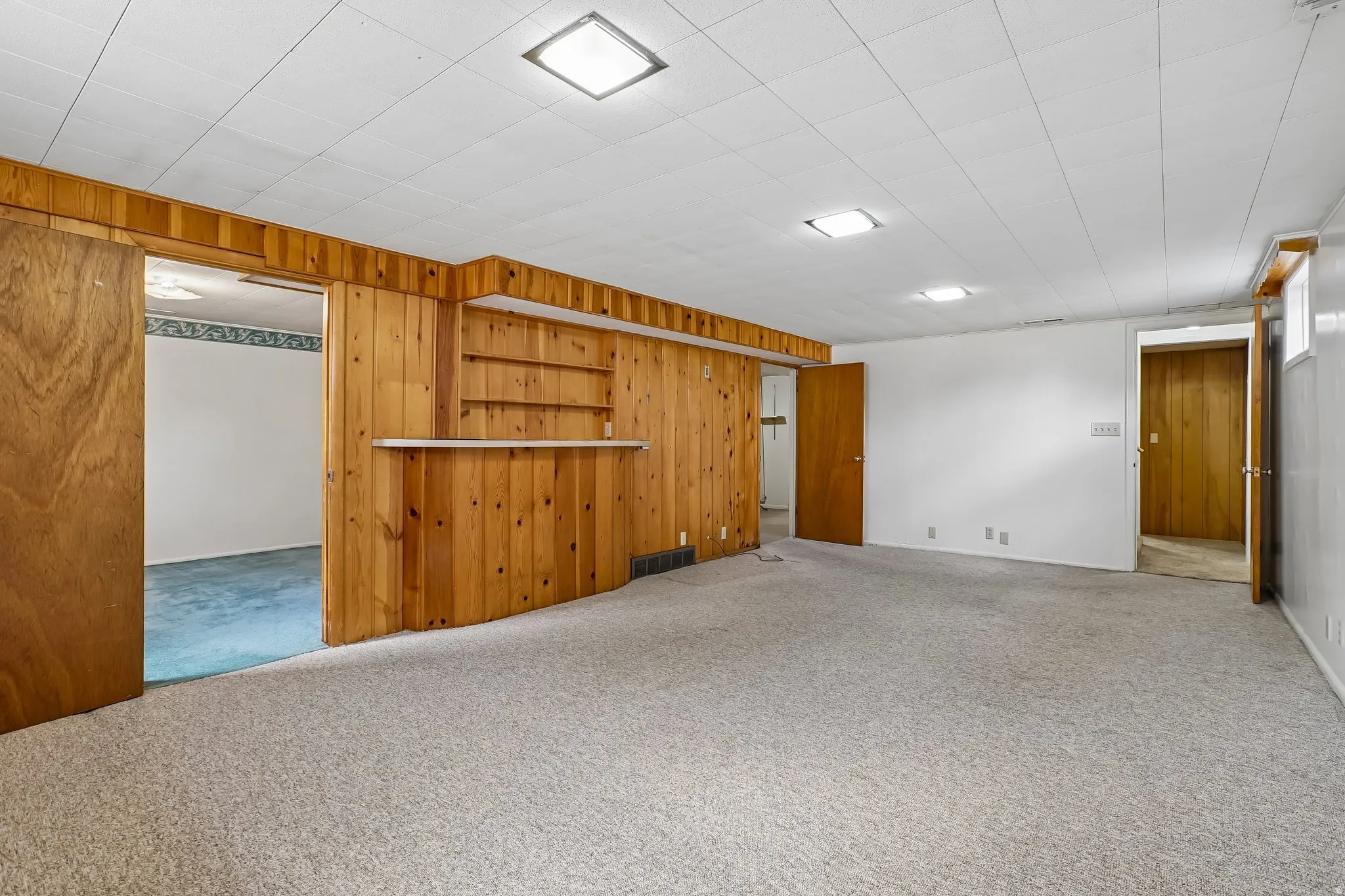 Spacious lower-level room featuring light gray carpeting, white walls, and a wood-paneled accent wall with built-in shelving and counter space