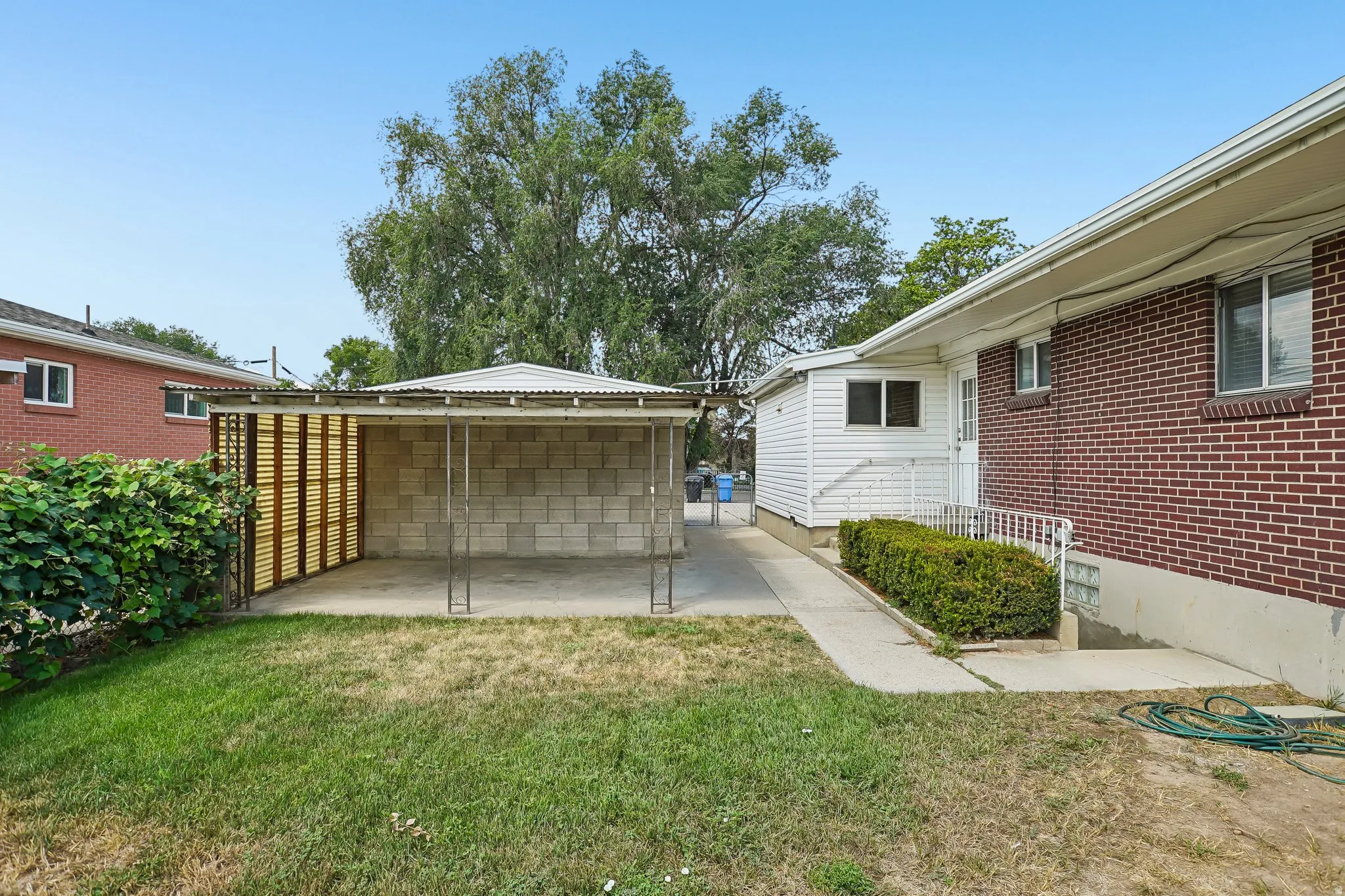 Expansive backyard featuring a concrete block carport with a corrugated metal roof, a secondary structure with white siding, a red brick exterior on the main residence, and a concrete pathway leading to an entry
